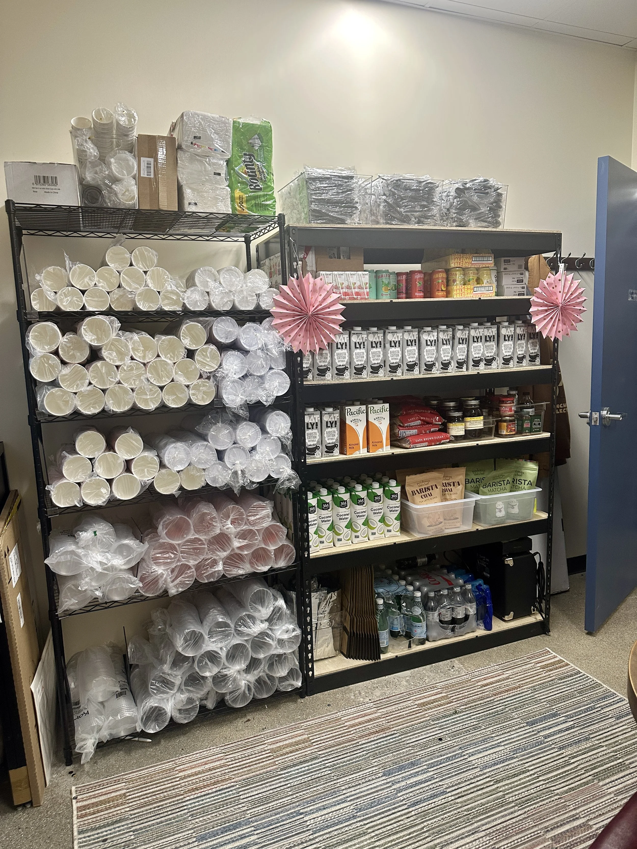Shelves stocked with disposable cups, paper bowls, paper towels, juice boxes, coffee cartons, and bottled water, with pink paper decorations on the sides.