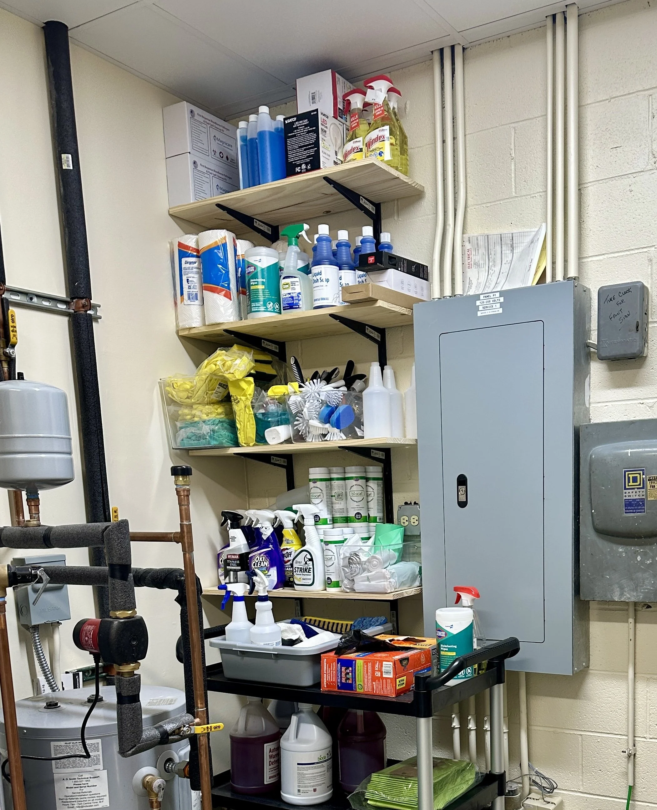 Shelves with cleaning supplies and tools in a utility room, including spray bottles, disinfectants, paper towels, and gloves.