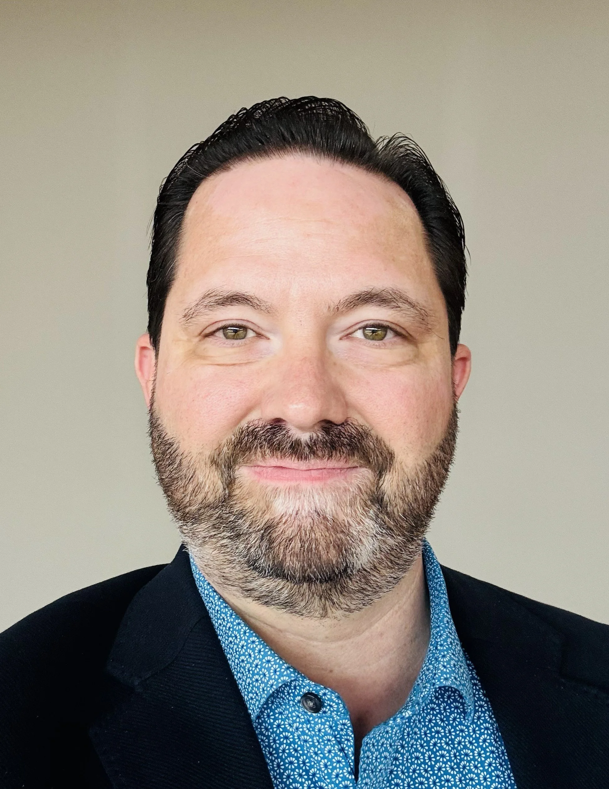 Close-up of a smiling man with dark hair, a beard, and hazel eyes, wearing a blue patterned shirt and a black jacket, against a plain background.