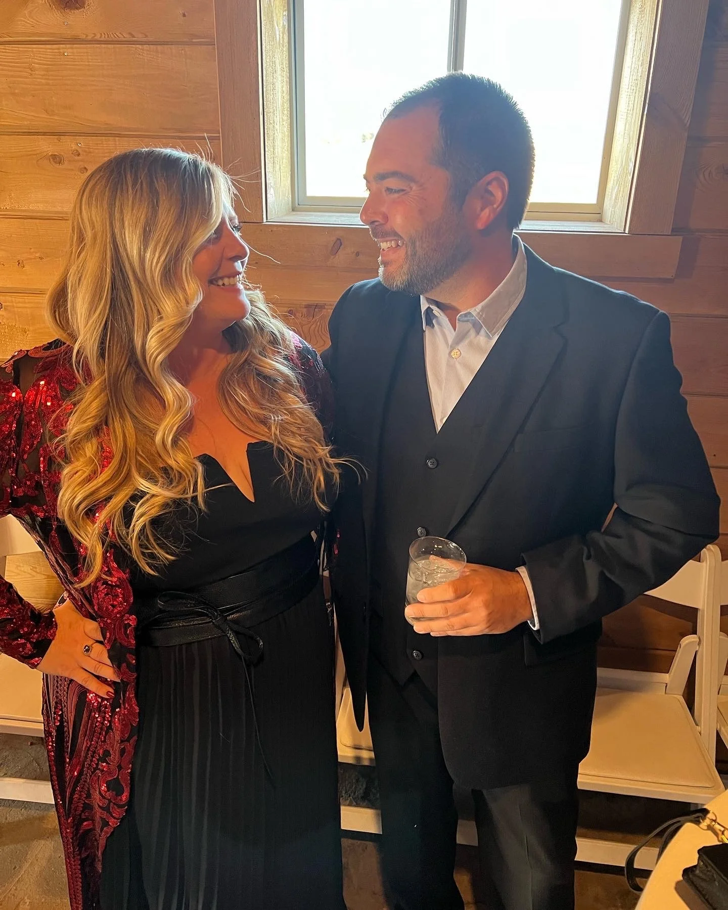 A man and woman are smiling and looking at each other in a warmly lit room with wooden walls and a window behind them. The man is wearing a dark suit and holding a drink, while the woman is dressed in a black and red outfit with long, wavy blonde hair.