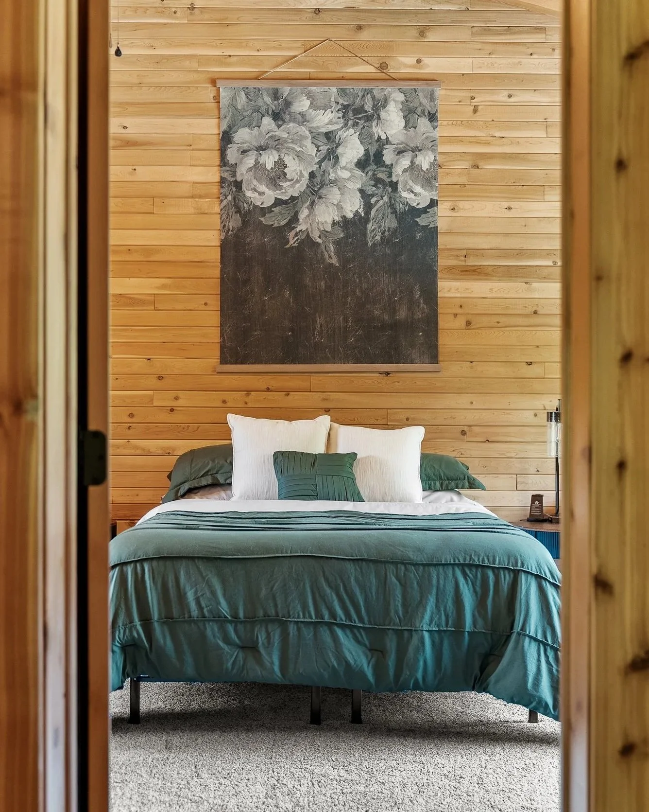 A neatly made bed with white and green pillows, covered with a green quilt, in a rustic bedroom with wood-paneled walls and a large floral painting.