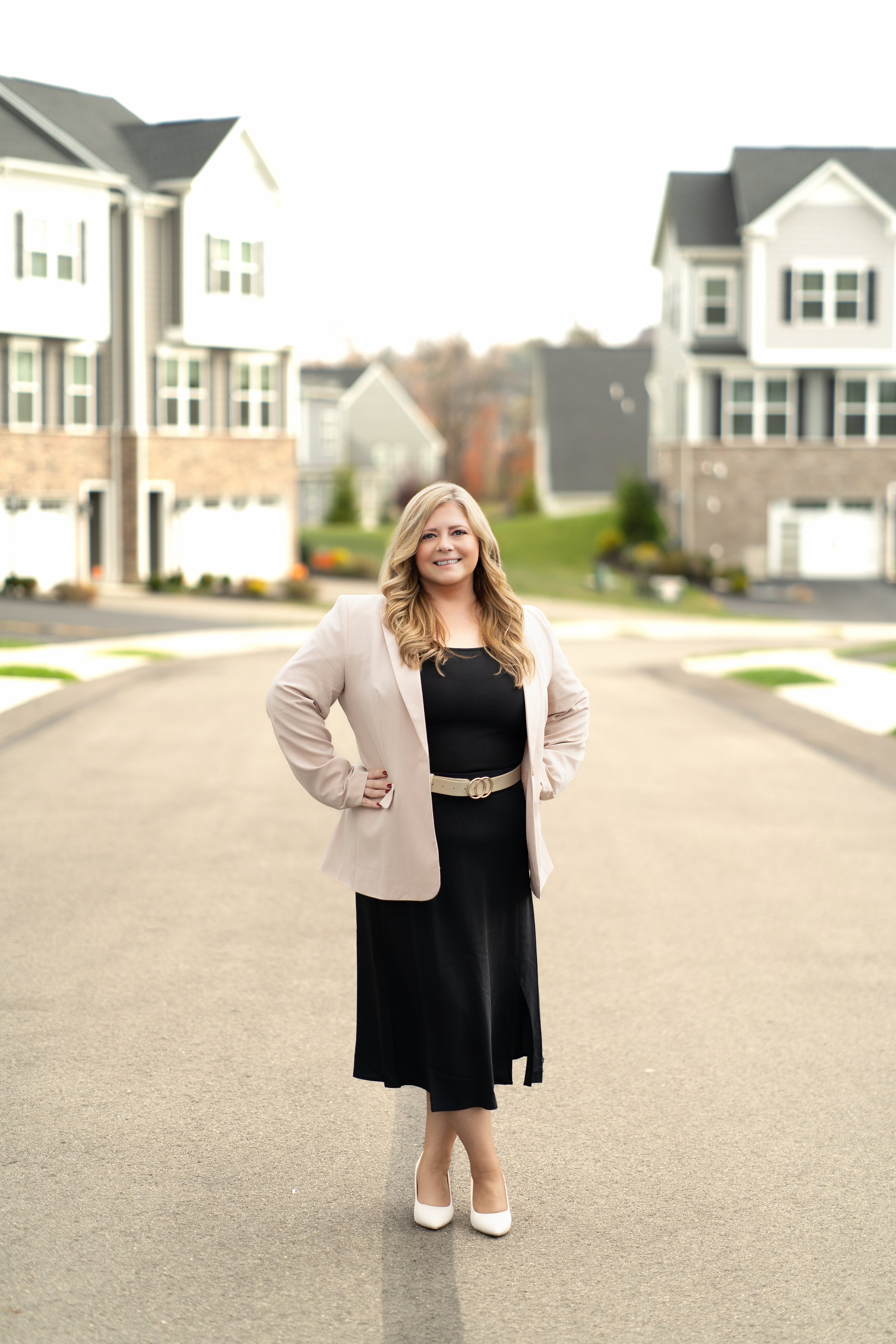 Professional portrait of Elissa Moore, Pittsburgh REALTOR® standing in the middle of a residential street, smiling, with modern suburban houses in the background. She is wearing a beige blazer, black dress, white shoes, and has long wavy blonde hair.