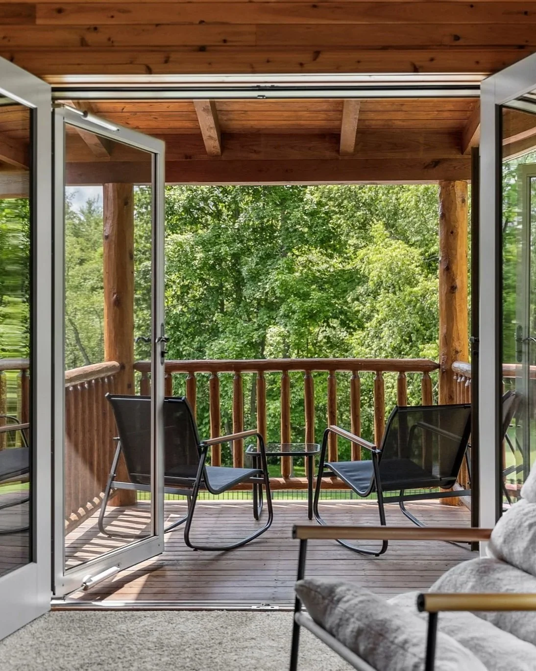 View from inside a cozy living room opening to a wooden balcony with chairs and trees in the background.