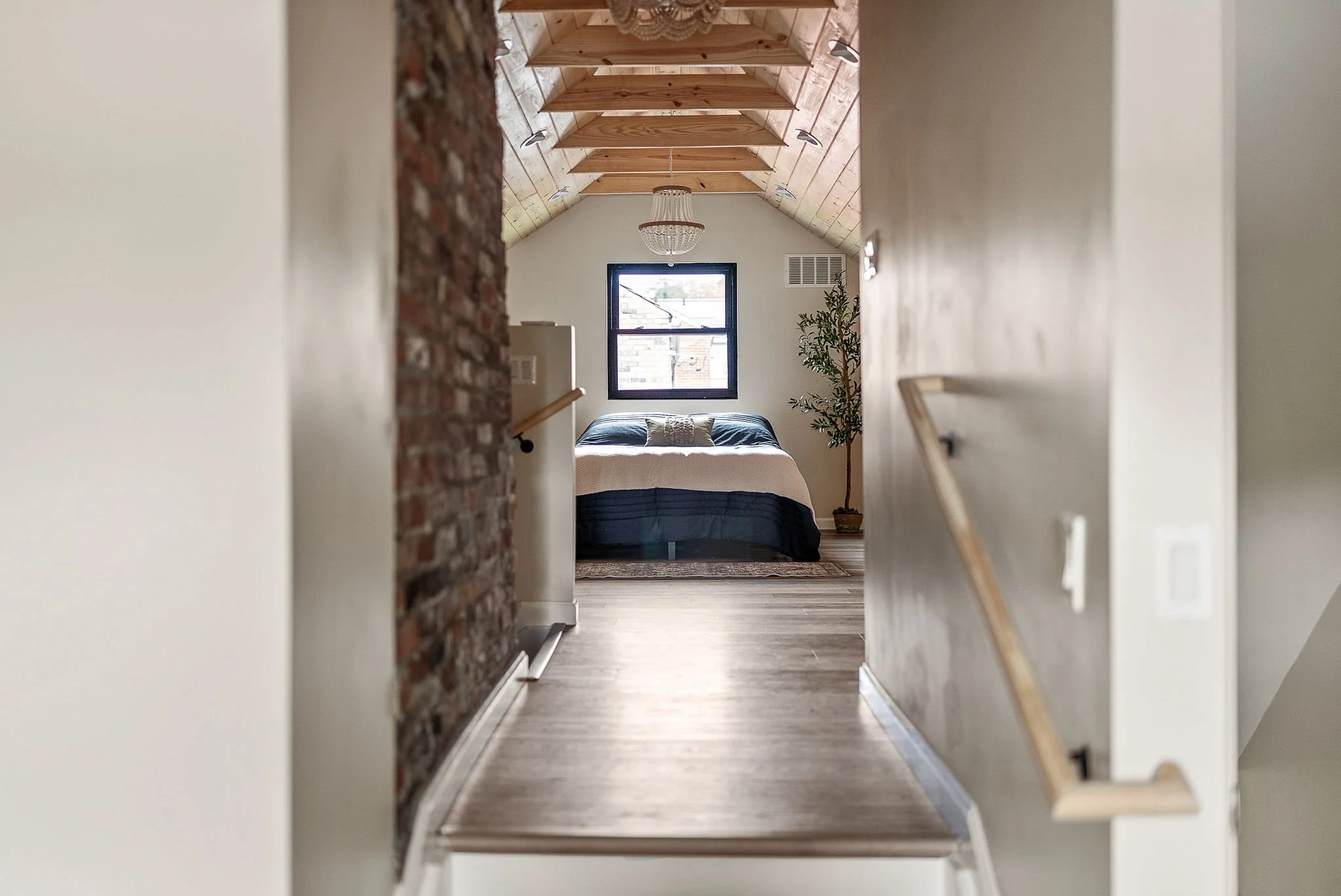 View of a cozy bedroom with a high wooden ceiling, a chandelier, a window, a bed with pillows, and a nearby potted plant, seen through a hallway with white walls and a wooden floor. representing the home selling journey in Greater Pittsburgh.
