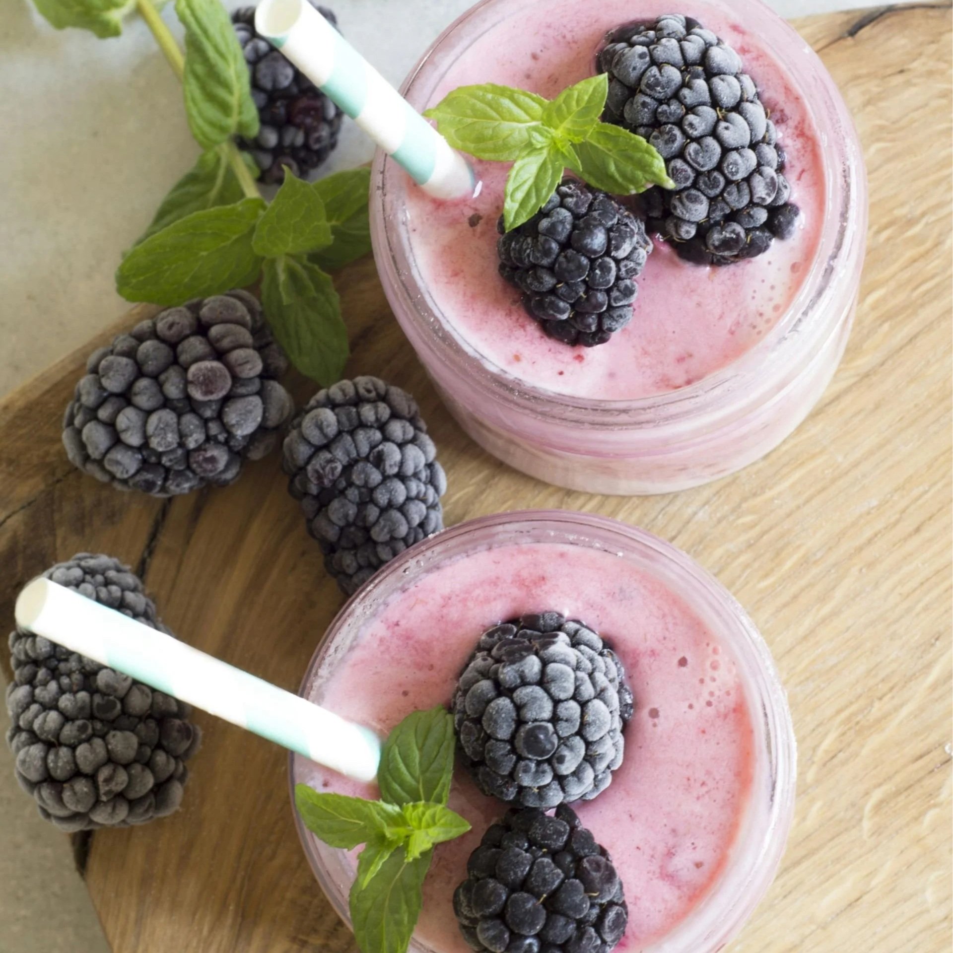 Two smoothies topped with blackberries and mint leaves, with additional blackberries and mint leaves on a wooden surface.