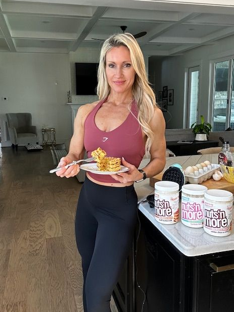 A woman with long blonde hair, wearing a pink sports bra and dark leggings, holding a fork and a plate of pancakes in a modern kitchen.