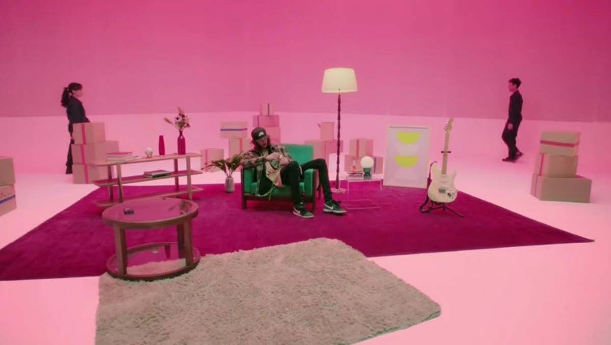 A person sitting in a green armchair in a pink-room, surrounded by modern furniture, a guitar, and standing individuals in the background.
