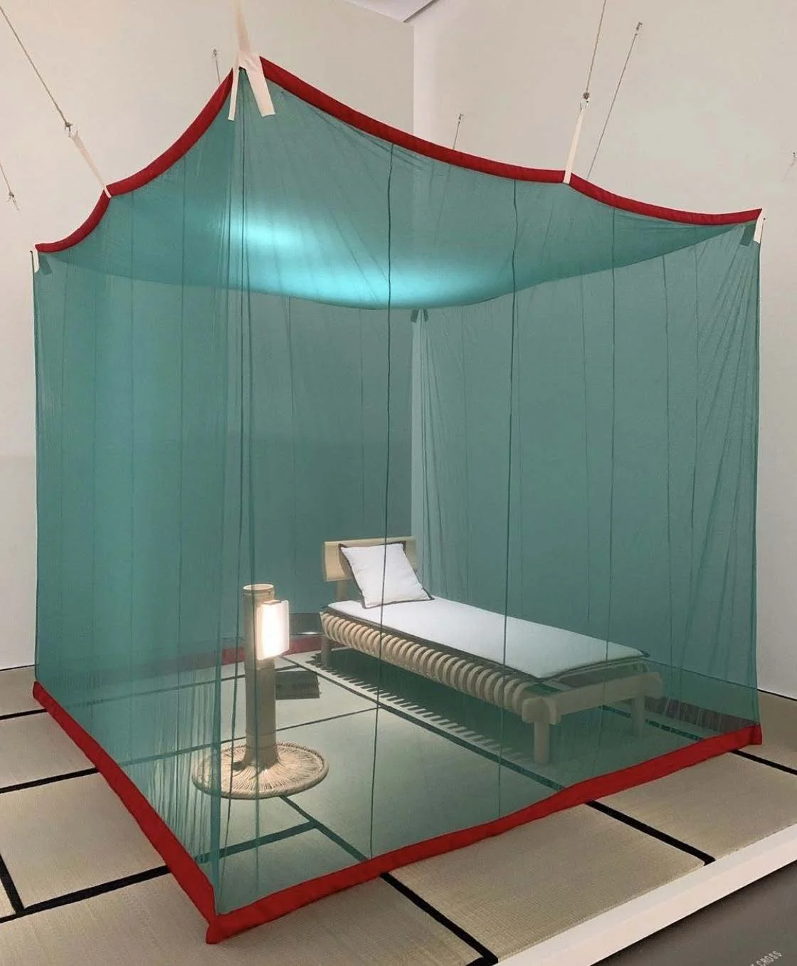 A bed inside a room enclosed by a green mosquito net with a red border, over a tatami mat floor. There is a small table with a lamp next to the bed.