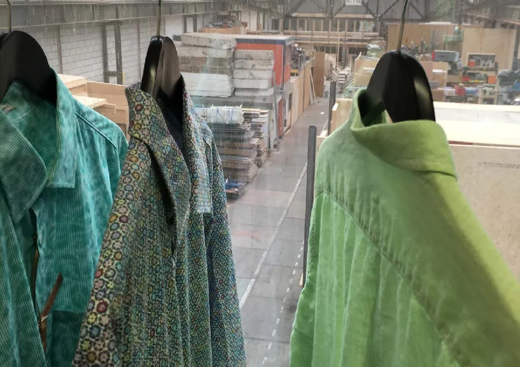Four jackets and coats displayed on black mannequin busts in a clothing store window, with a background of a workshop or storage area with shelves and boxes.