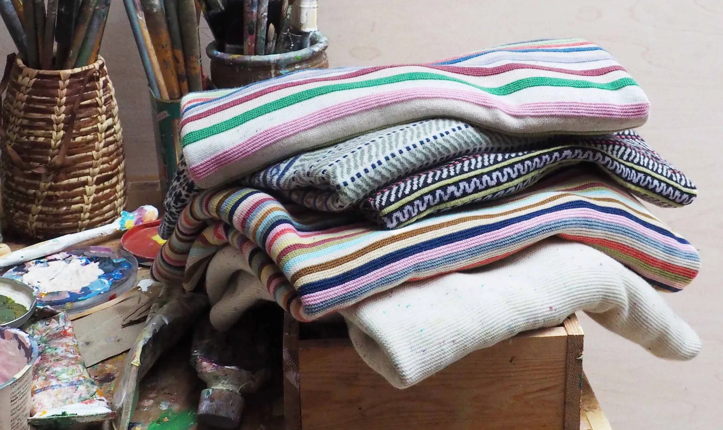 Stack of striped Mansted knitwear beside an artist's paint palette and brushes