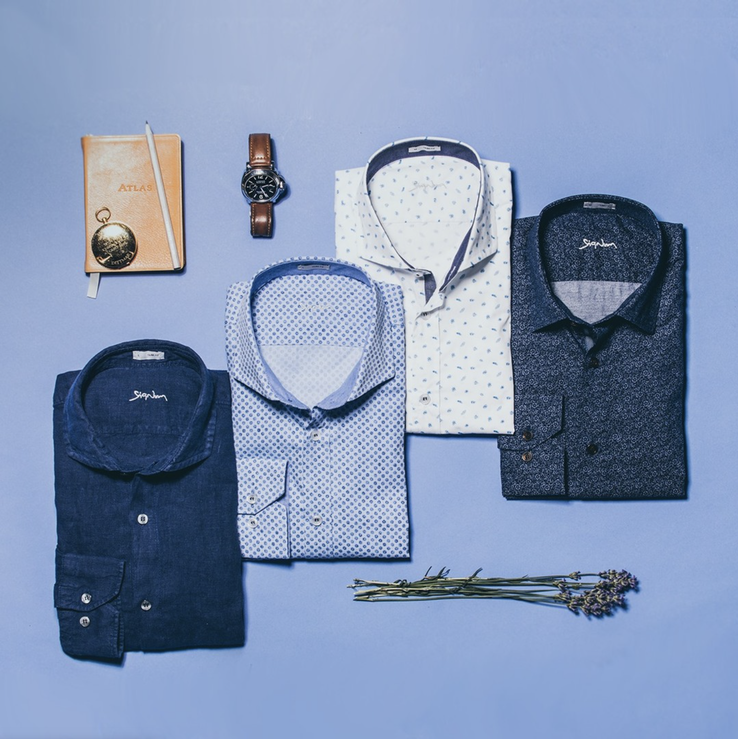 Flatlay of folded Signum dress shirts with a wristwatch, notebook, and lavender sprig on a blue background