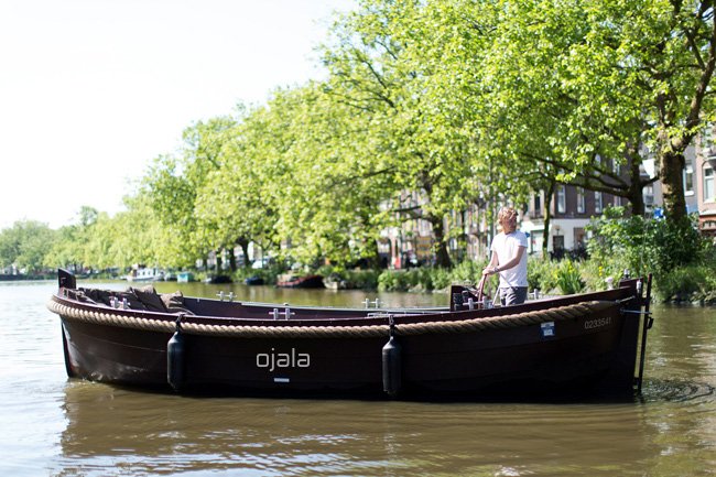A person standing on a black boat named 'ojala' on a canal with green trees and buildings in the background.