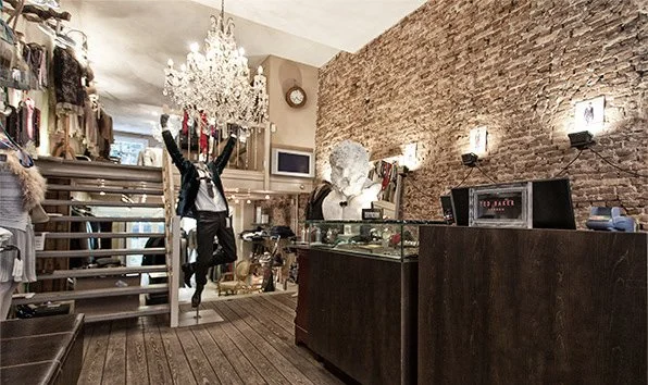 Interior of a clothing store with a chandelier, brick wall, staircase, and a mannequin with raised arms.