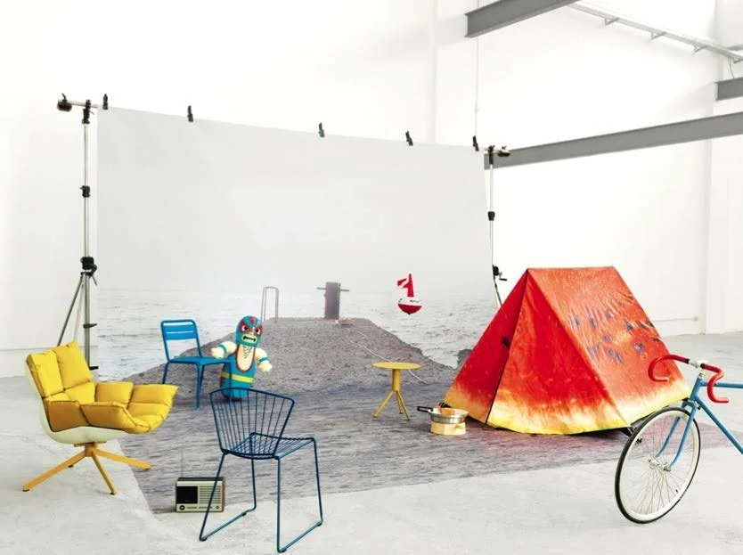 Indoor scene with a beach backdrop, yellow armchair, blue chair, wire chair, small table, tent, bicycle, and a puppet figure, creating a cozy, eclectic setup.
