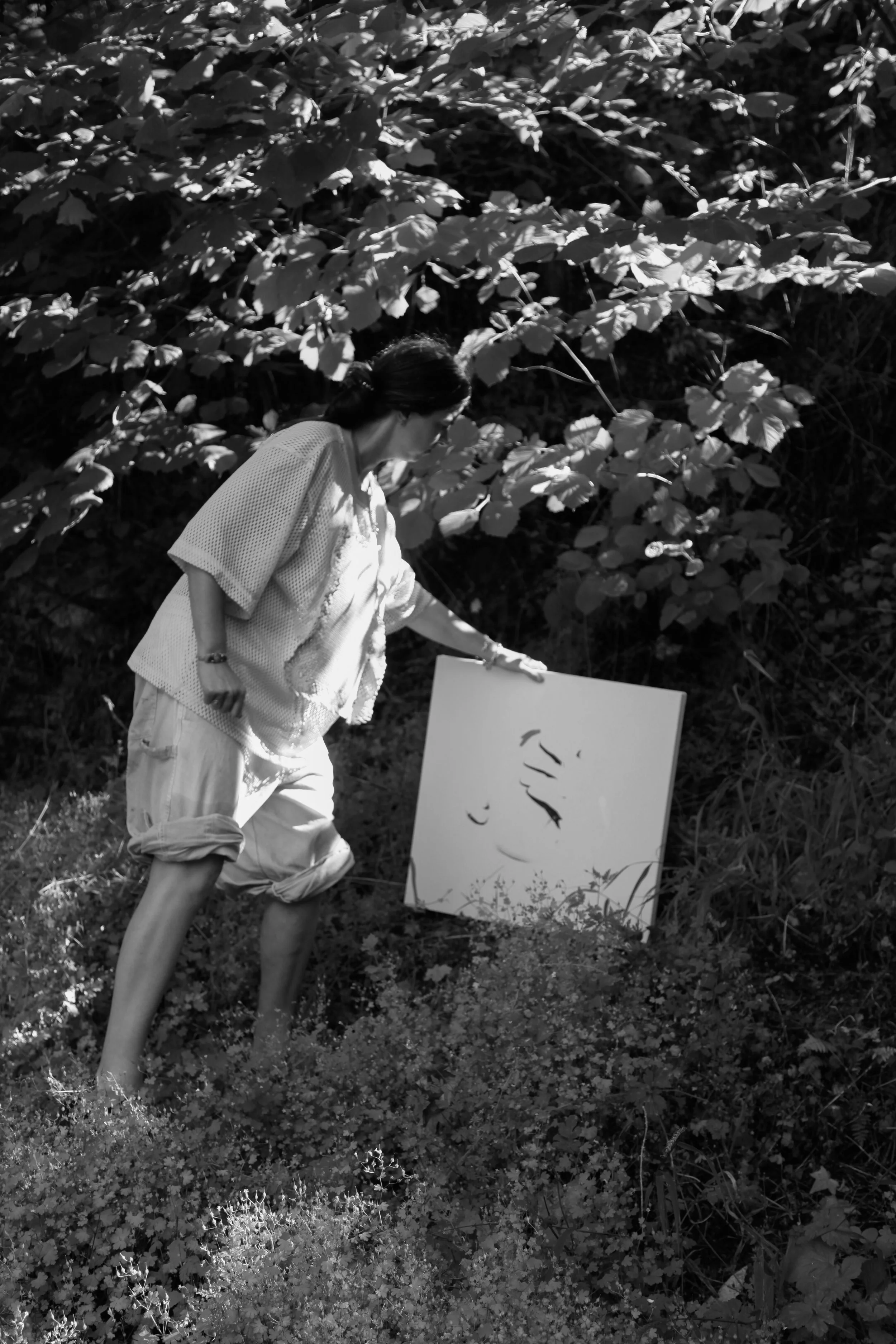 A woman with dark hair tied back, wearing a light-colored kimono and loose pants, is outdoors among bushes and small plants, holding a white canvas with an abstract black ink drawing, with her head turned to the side.