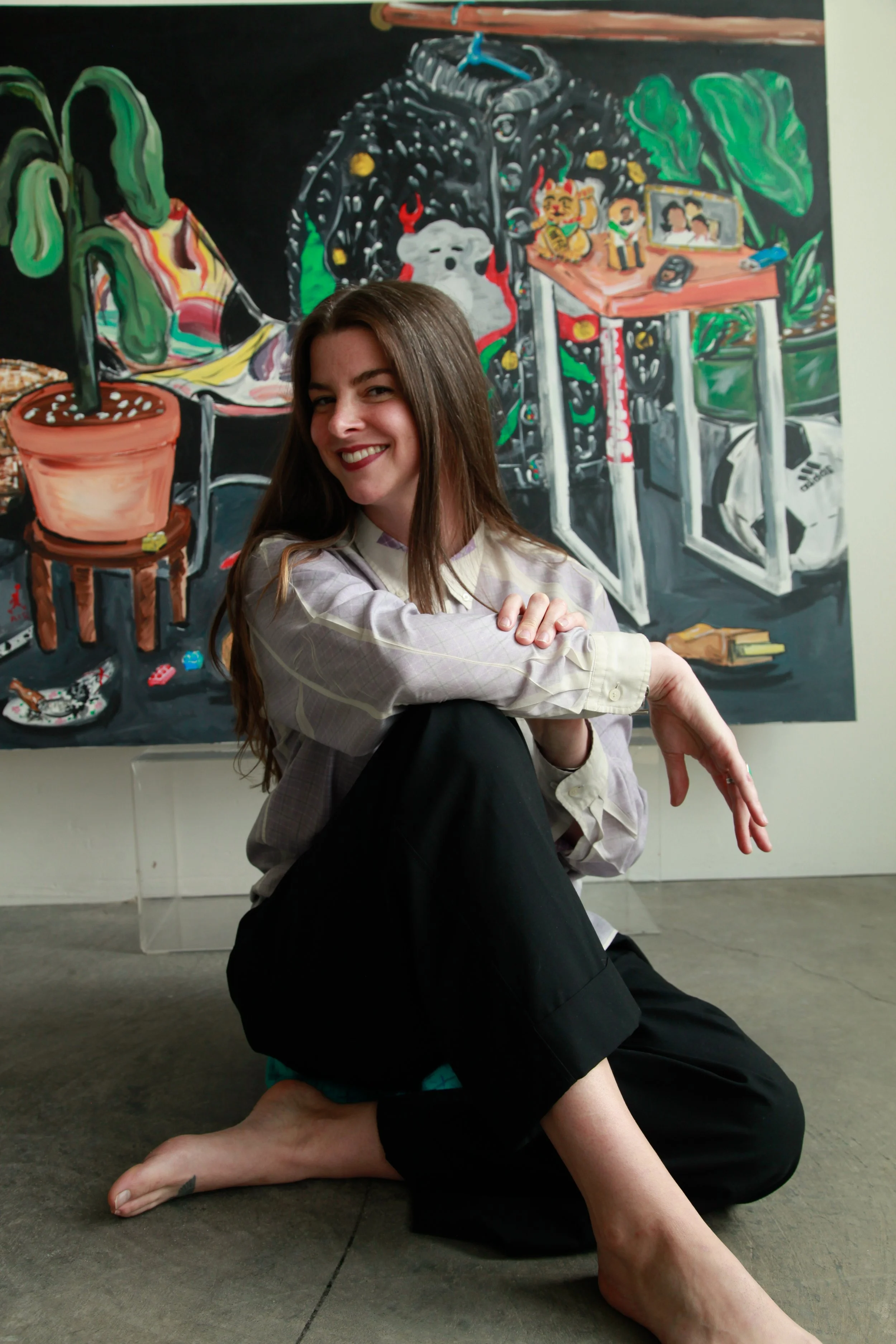 A woman with long brown hair smiling, sitting on the floor with one knee bent and her arms crossed over her knees, in front of a colorful abstract painting. The painting features various figures and objects like a dog, a table with books, green plants, and other artistic elements.