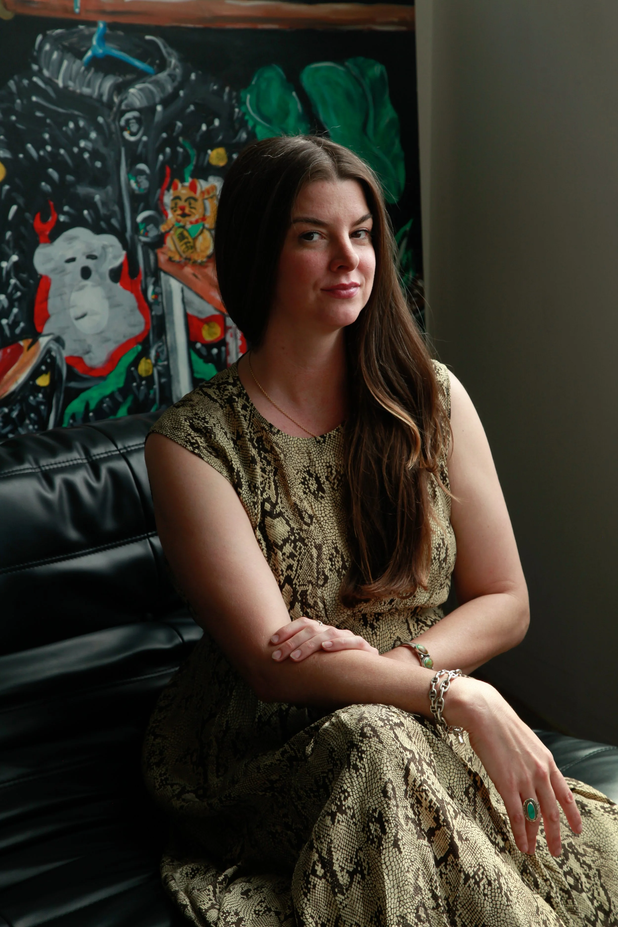 A woman with long brown hair sitting on a black leather bench, wearing a snakeskin patterned dress and jewelry, with colorful artwork in the background.