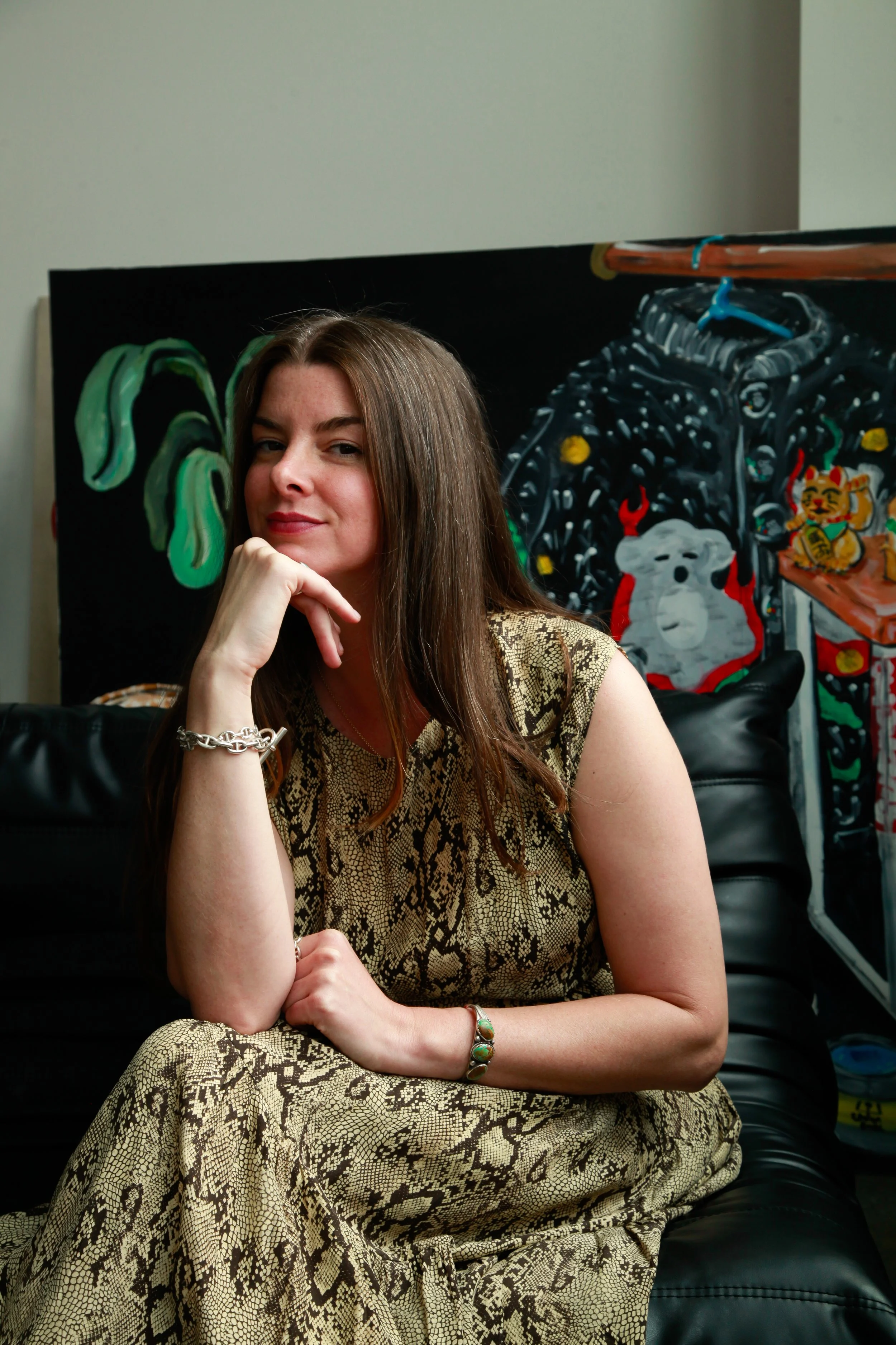 A woman sitting on a black leather chair, wearing a snake print dress, with her left hand resting on her chin. Behind her, there's a colorful painting of a dog, a tiger, and a motorcycle.