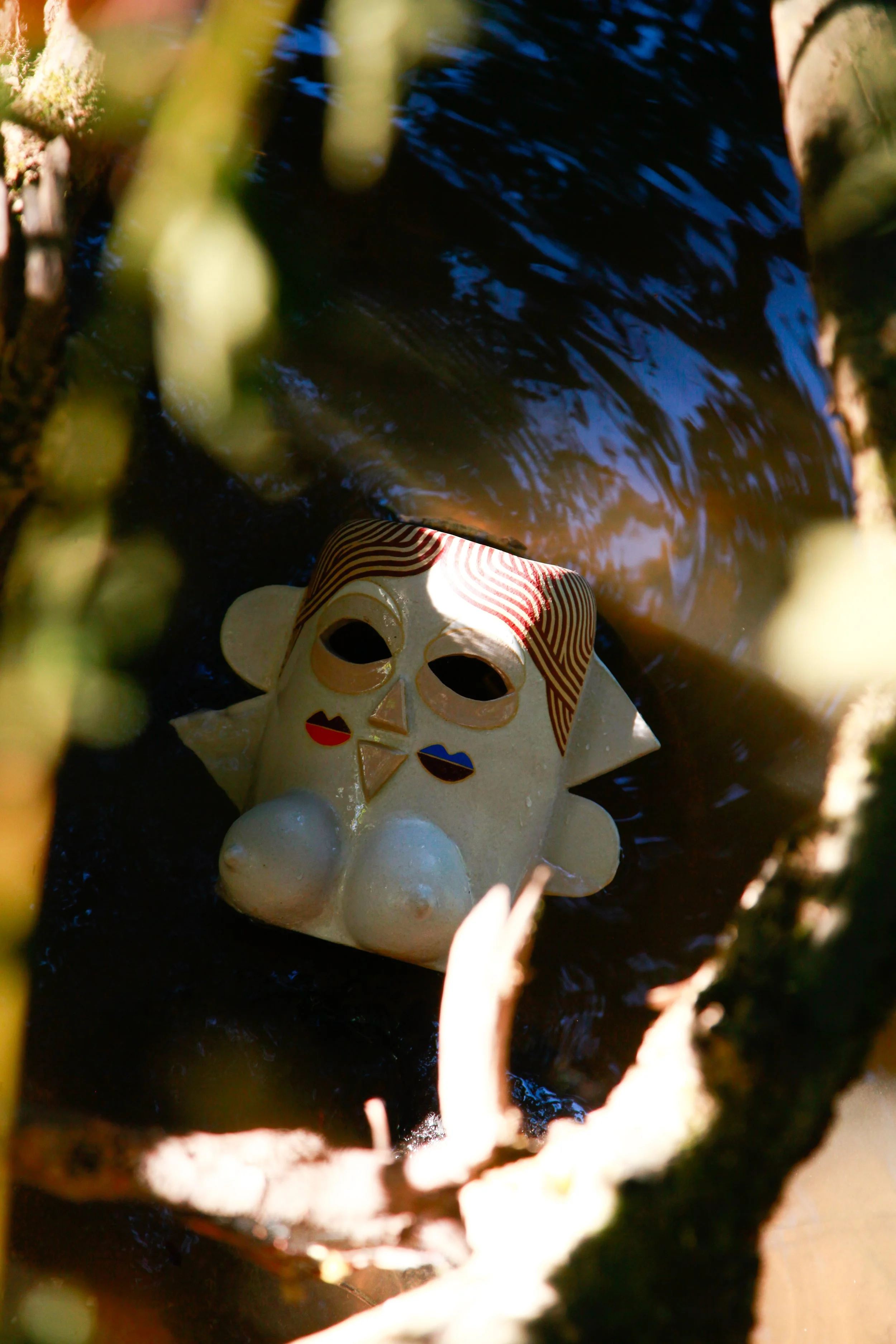 Colorful ceramic mask with a smiling face, hidden among green branches and leaves, partially submerged in water.