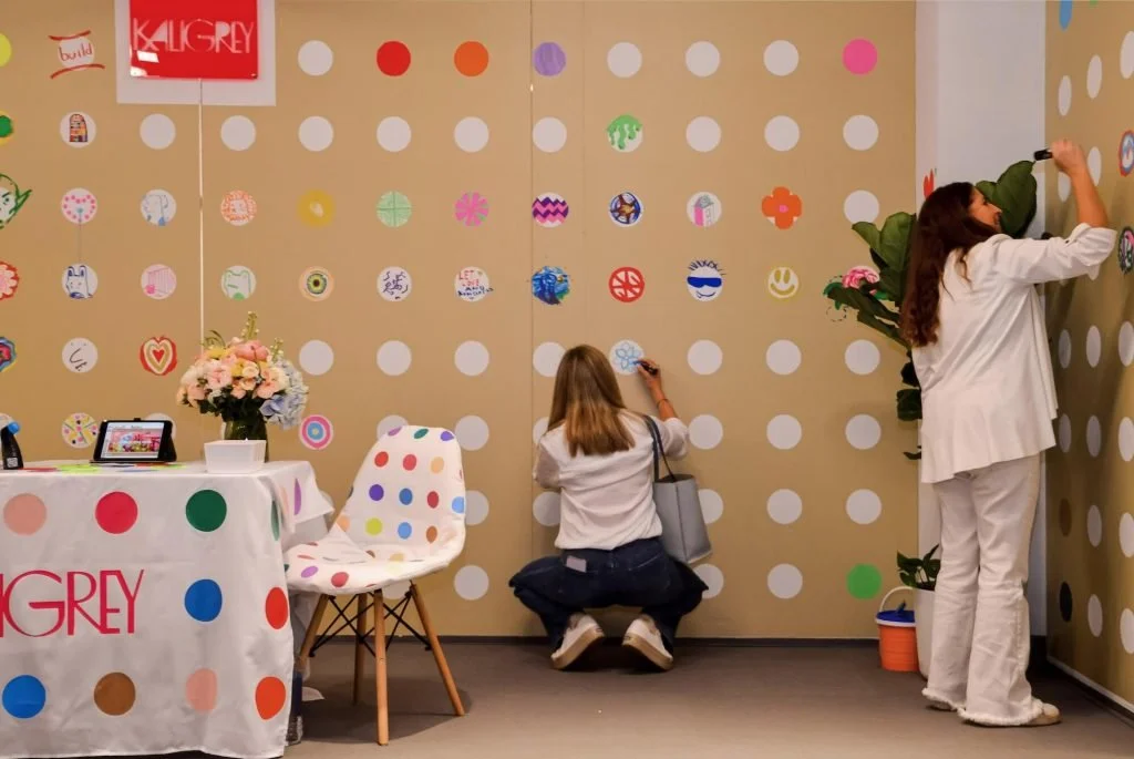 Two women are decorating a wall with colorful circular stickers, with a table holding a flower arrangement and a framed photo nearby.