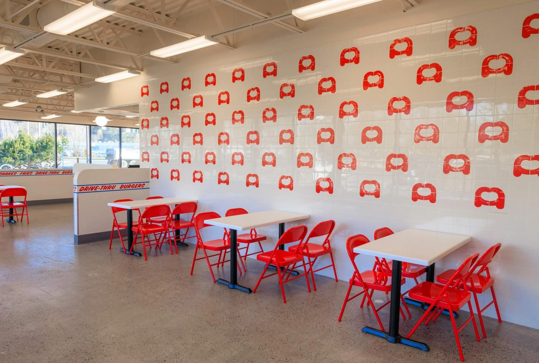 Interior of a fast food restaurant with white tiled walls decorated with red logos, red chairs, white tables, and large windows showing trees outside.