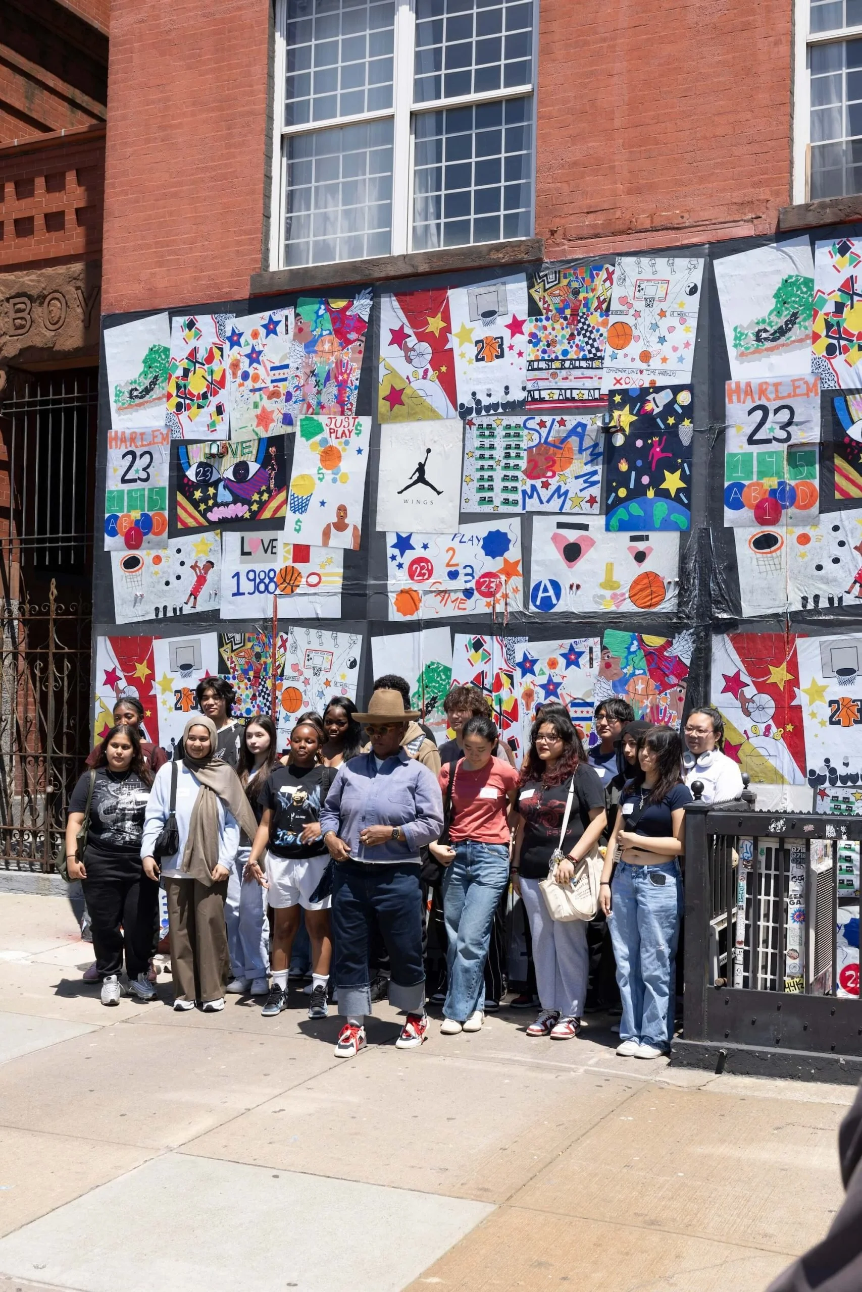 A group of diverse young people standing together on a street in front of colorful artwork displayed on a fence, with a red brick building and large windows in the background.