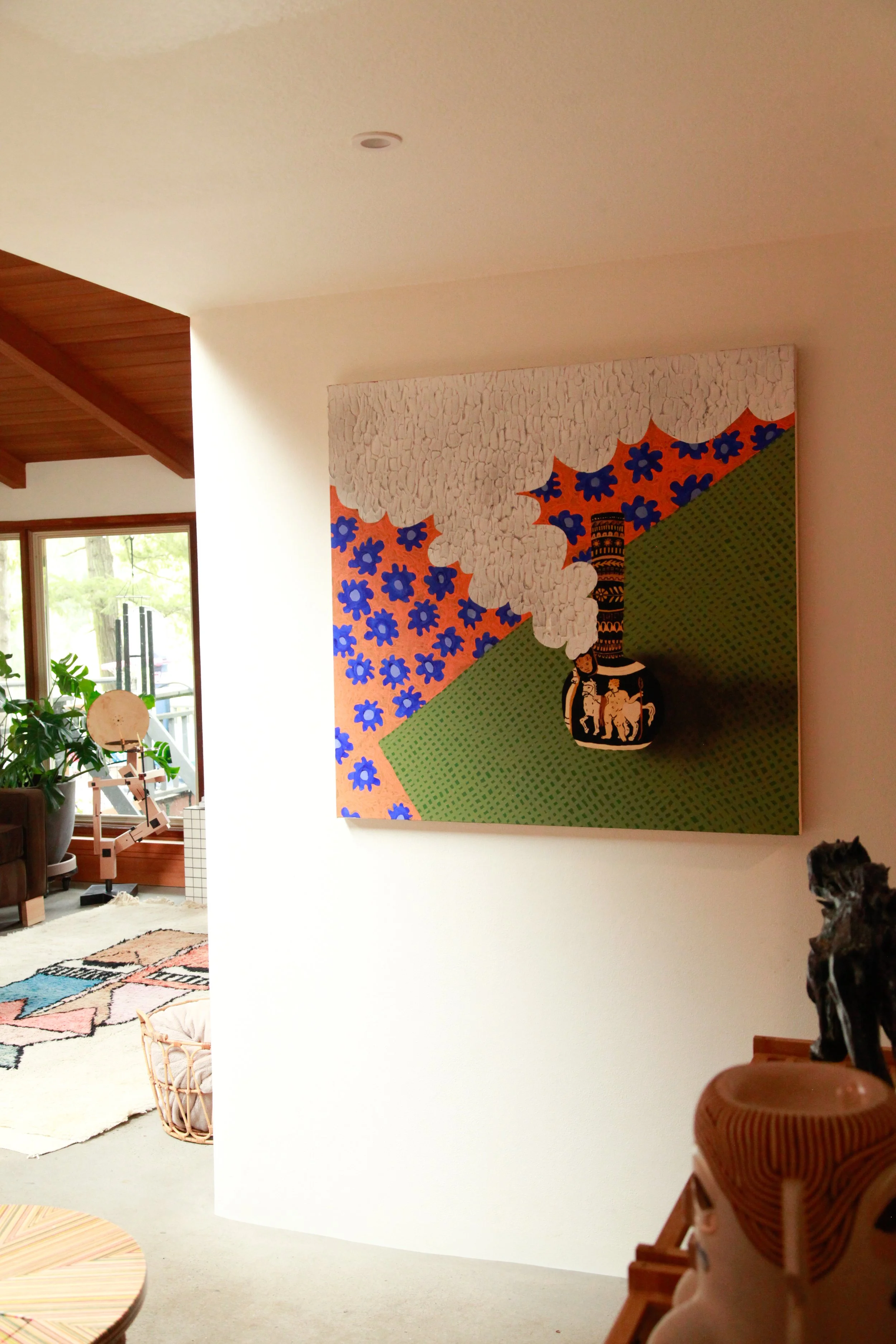 Part of an indoor living space showing a white wall with colorful artwork, a corner of a room with a window, a plant, a pattern rug, and a wooden piece with a figurine.