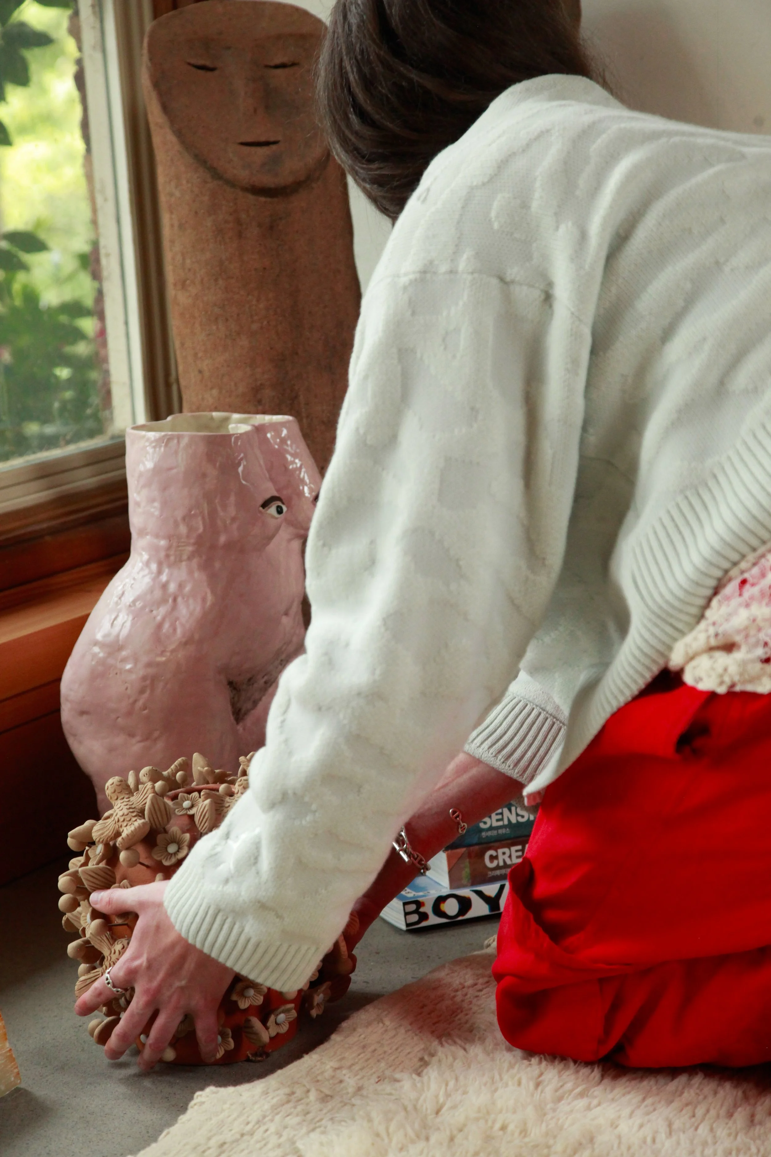 A person in a white sweater with rolled-up sleeves and red pants is arranging ceramic or clay art pieces on a surface near a window. Behind them, on the wall, is a tall, slender sculpture with a human face carved into it, and a pink ceramic vase with a face design is nearby. There are some boxes and art supplies around, and the scene is well-lit by natural light.