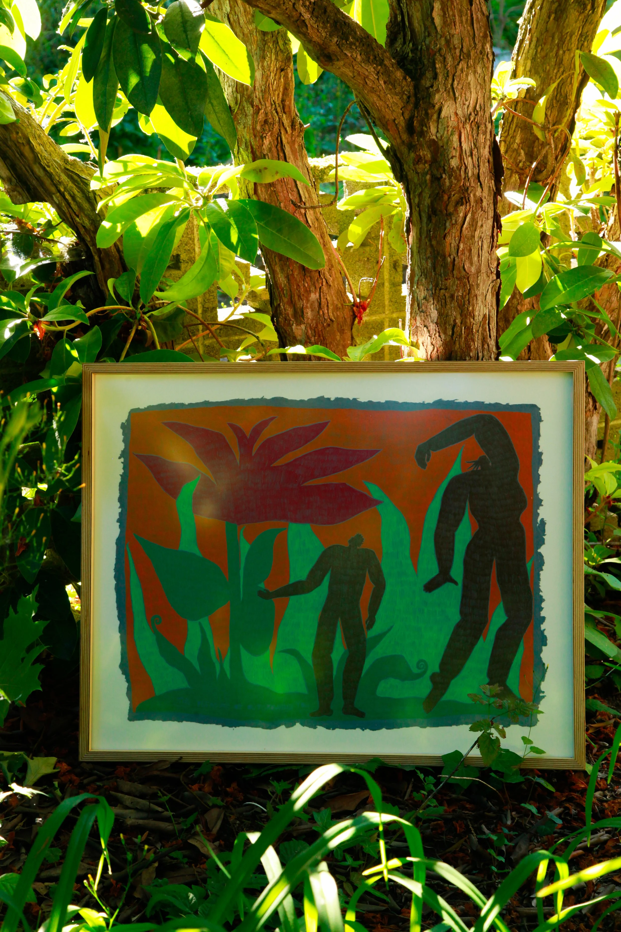 A framed painting propped up against a tree with green leaves, depicting silhouetted human figures among large stylized plants and a bright orange background.