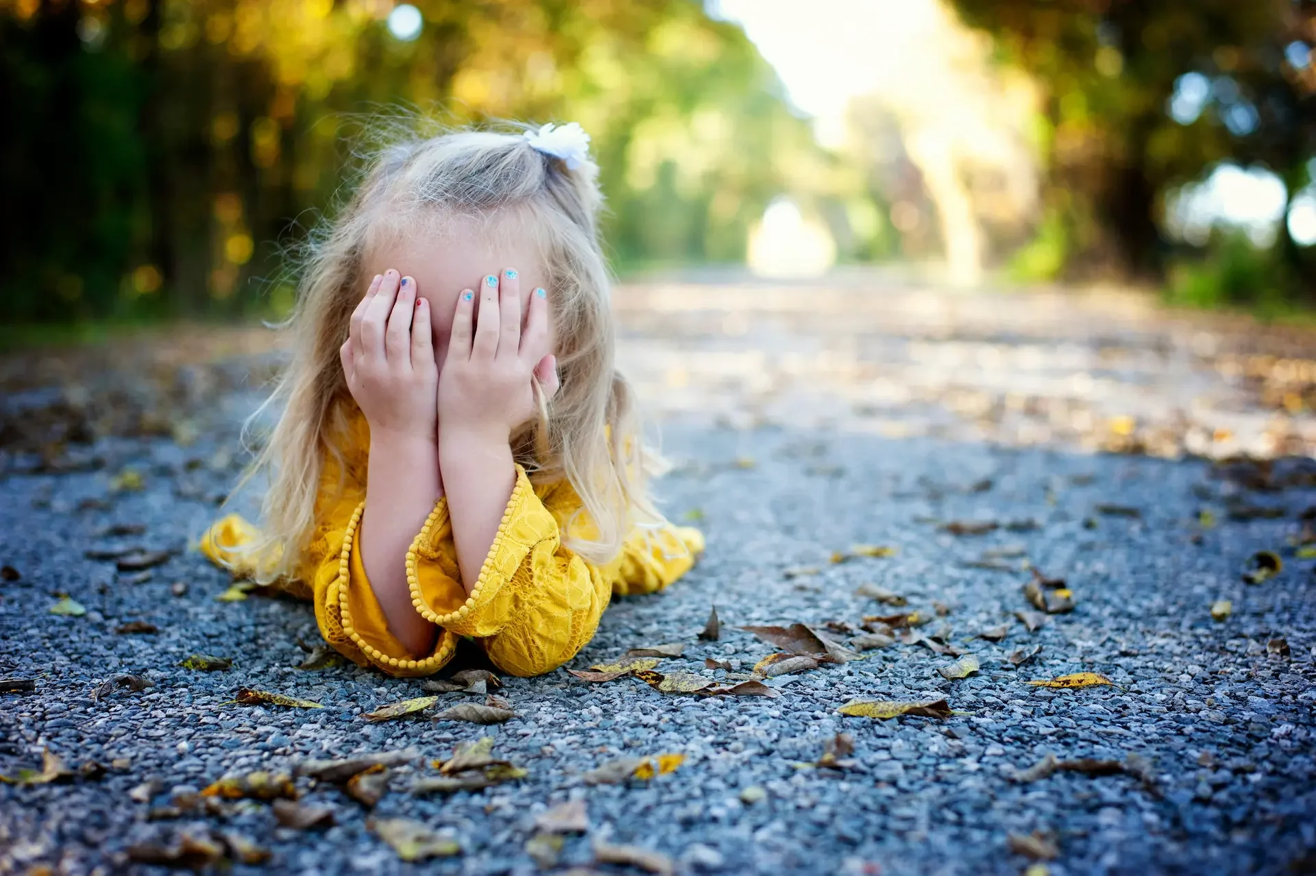 Little girl in yellow dress with hands in face, laying on ground - bright sunny