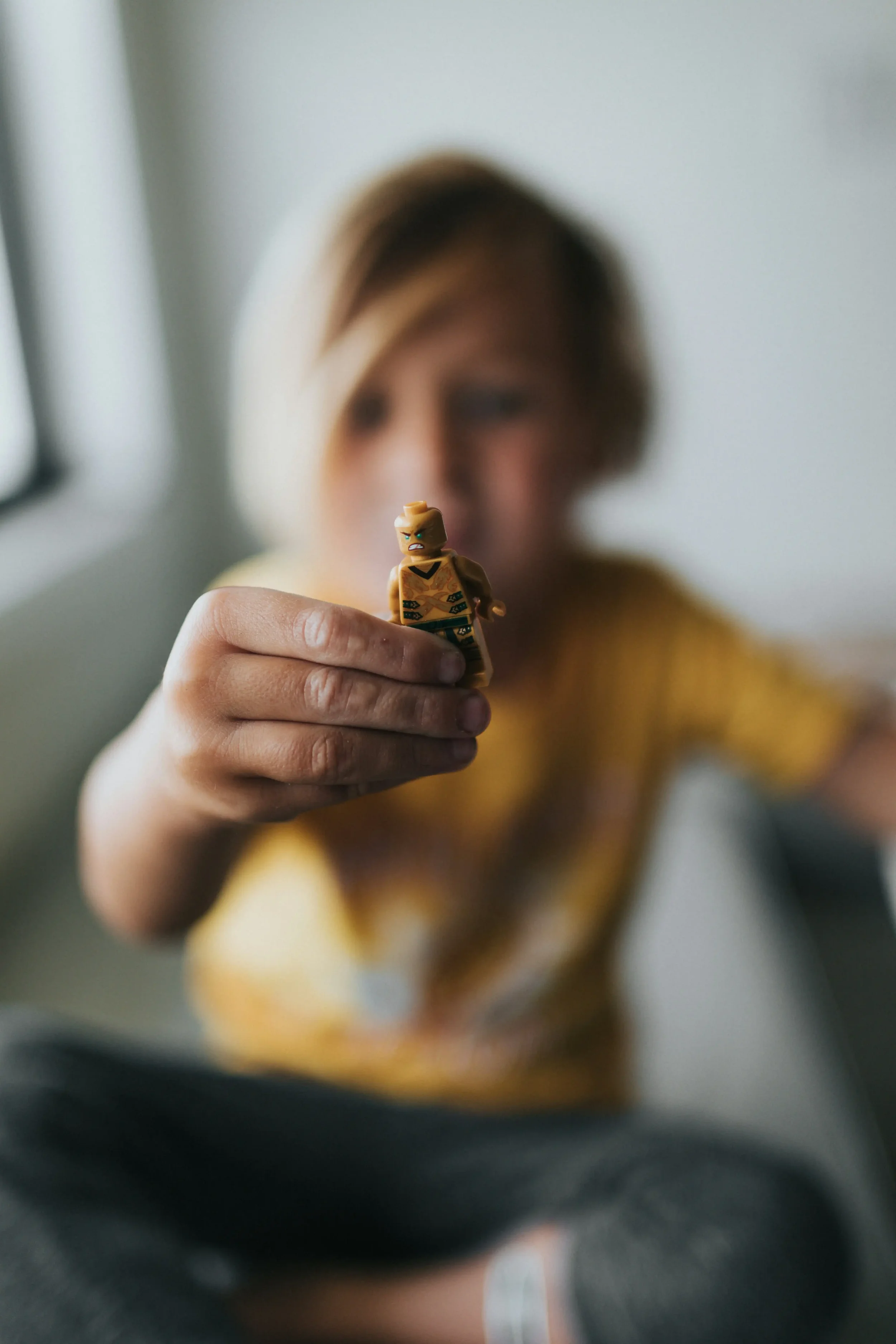 A child holding a LEGO minifigure with a grumpy expression in front of their face, with a blurred background.
