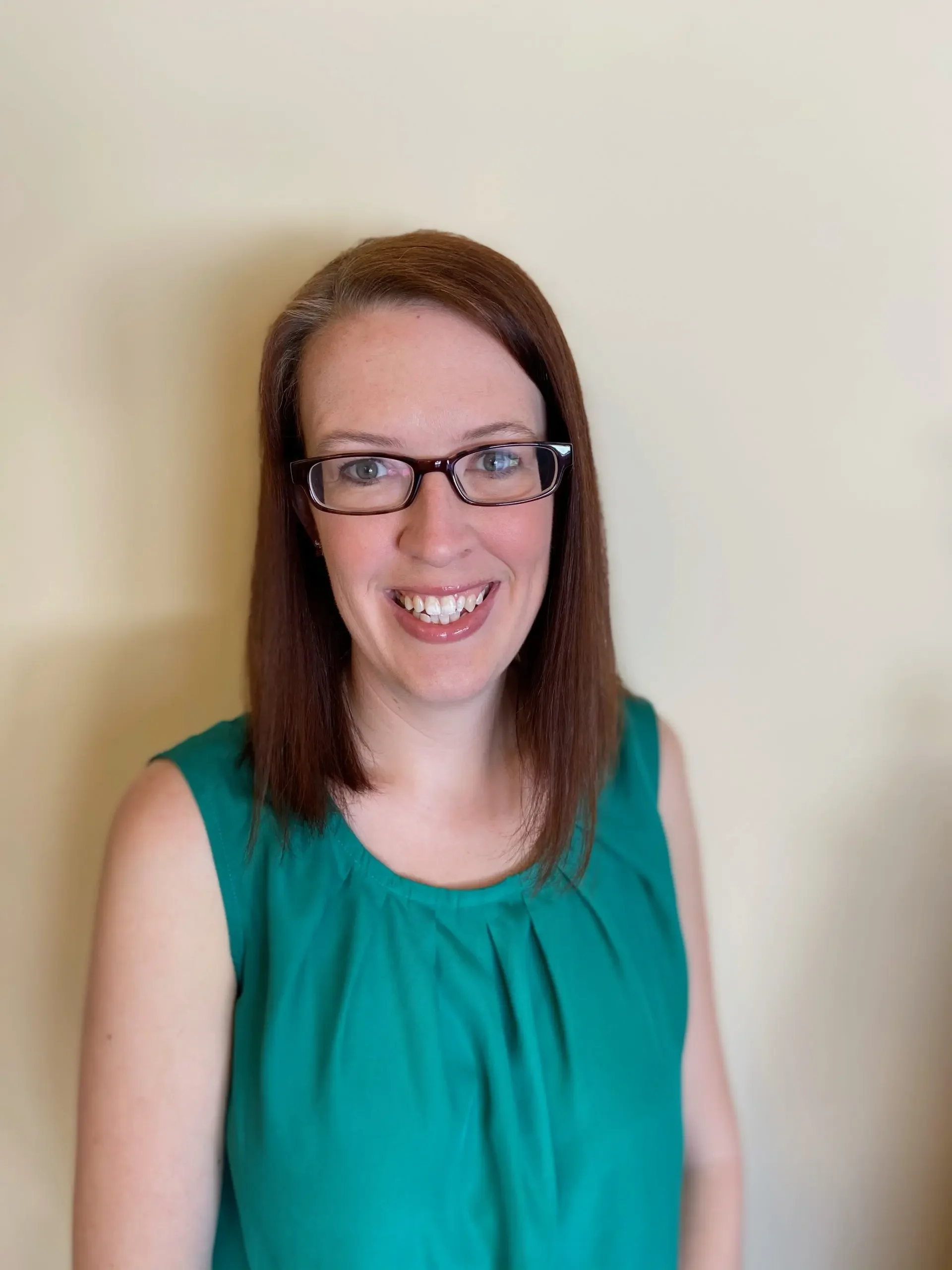 Erika Rodkey: Wearing glasses and a wide smile, wearing a teal top, standing against a plain light-colored wall. Therapist at Edith Schatz LLC