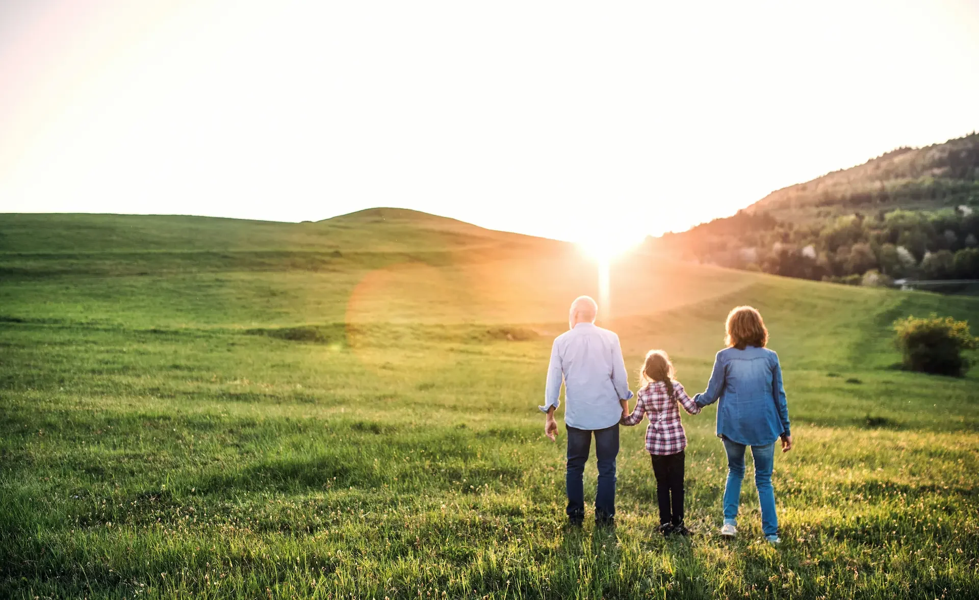 Three people, two parents and a child, walking hand in hand through a grassy field at sunset, with mountains in the background.