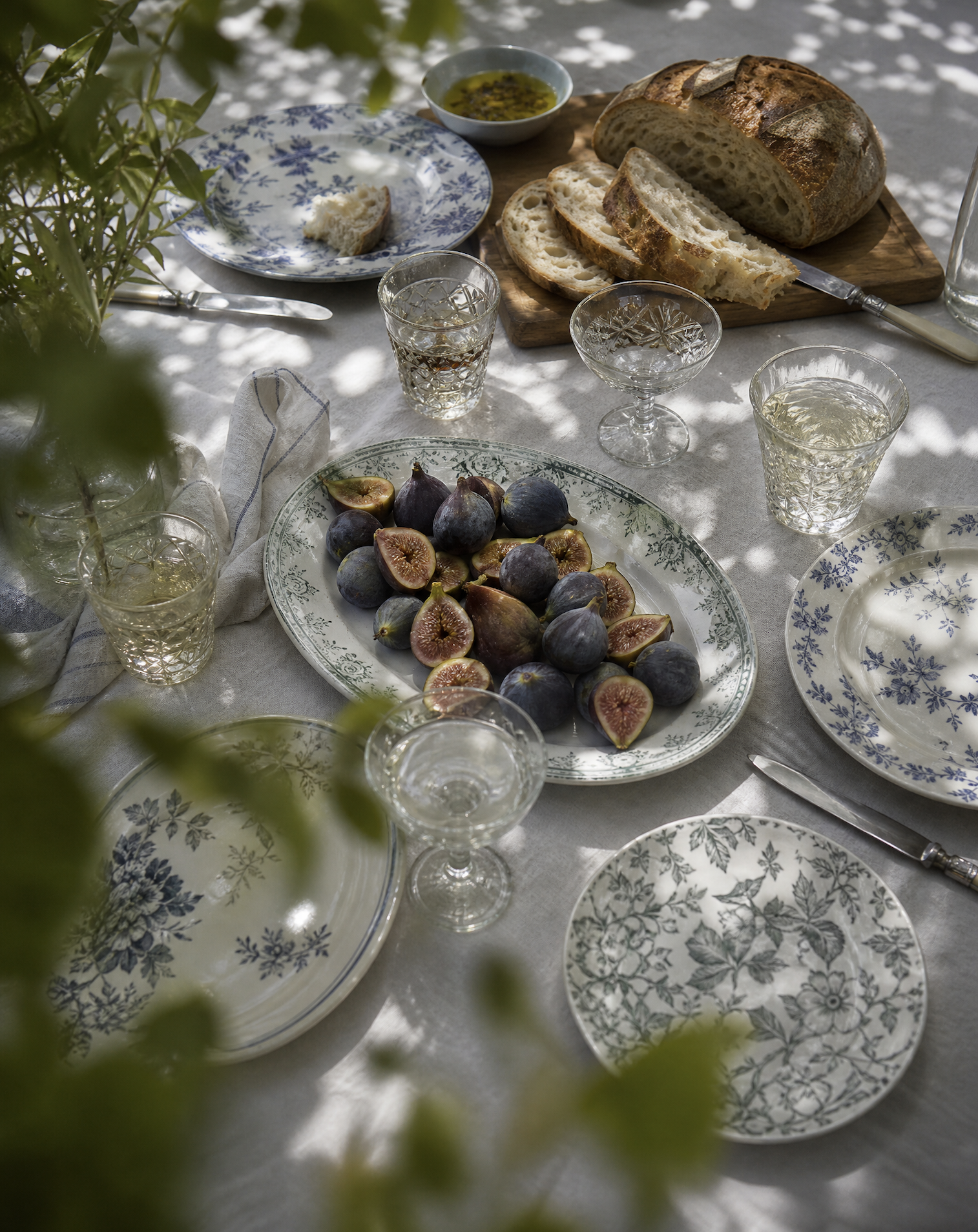 A table set for a meal with barley bread, figs, and glassware with a white tablecloth and blue and white patterned plates.