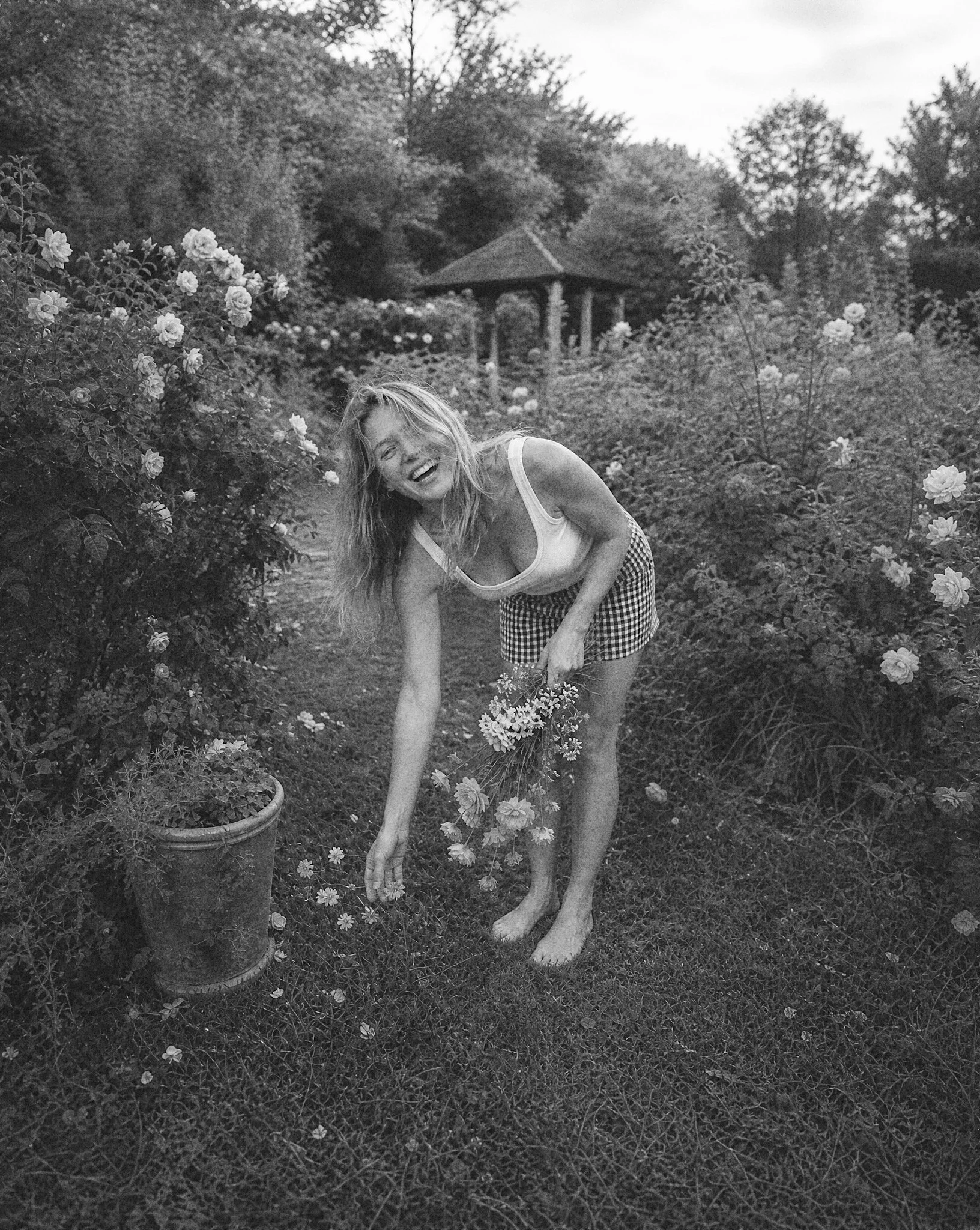 A woman with long hair, wearing a sleeveless top and checkered shorts, is smiling and bending over in a garden full of blooming flowers and shrubs. There are large flower pots nearby and a gazebo or pavilion in the background.
