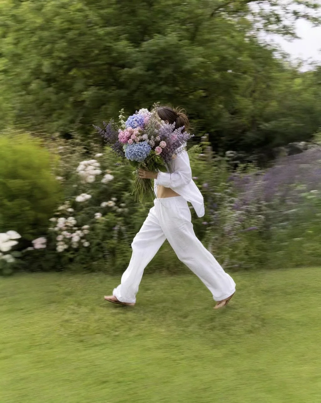 Person in white clothes walking on grass carrying a large bouquet of colorful flowers, with trees and shrubs in the background.