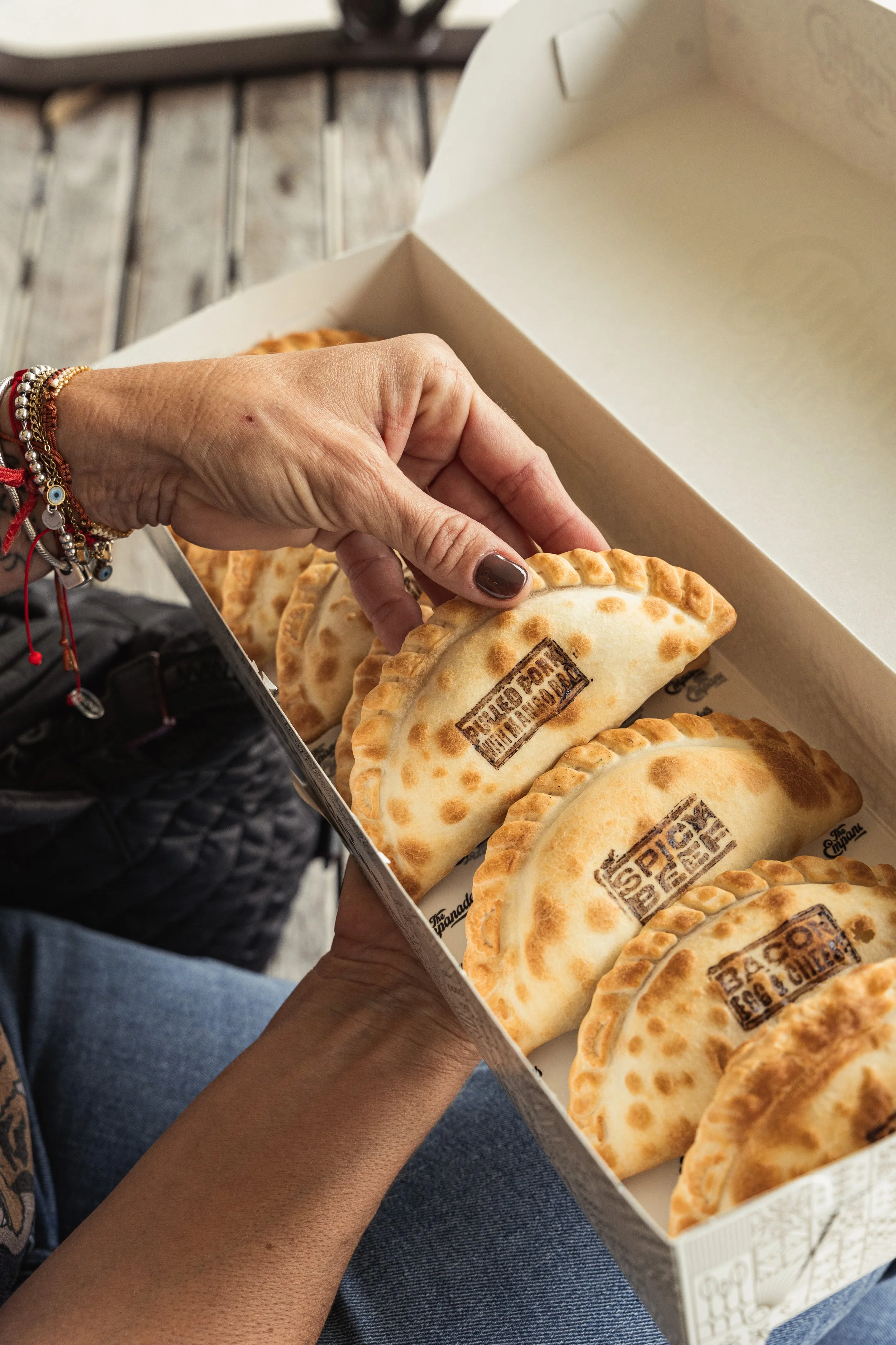 A person's hand with brown painted nails and multiple bracelets reaching into a box of baked empanadas with stamped logo, on a wooden surface.