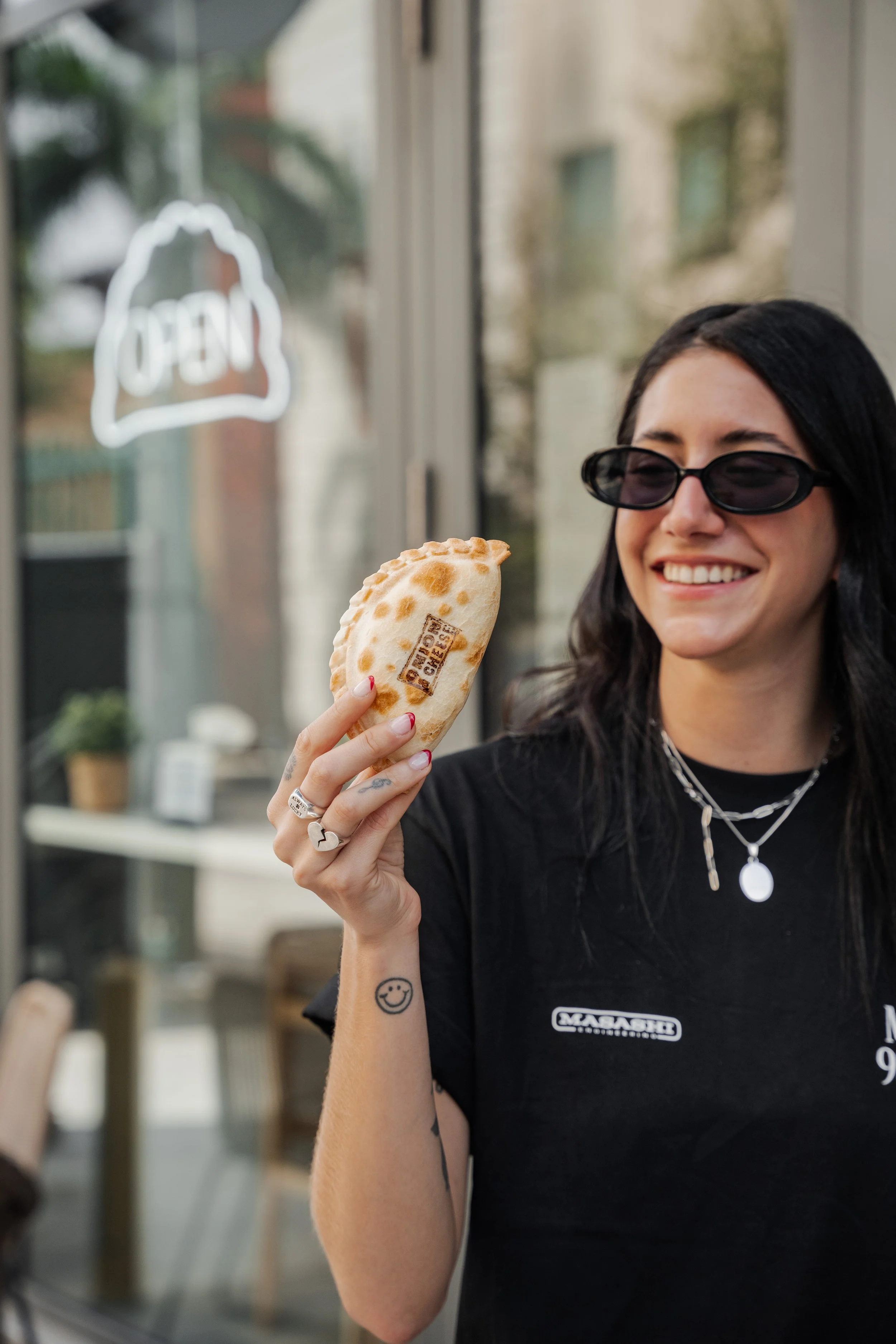 Woman holding a pepperoni calzone, smiling, wearing sunglasses, with a smiley face tattoo on her arm.