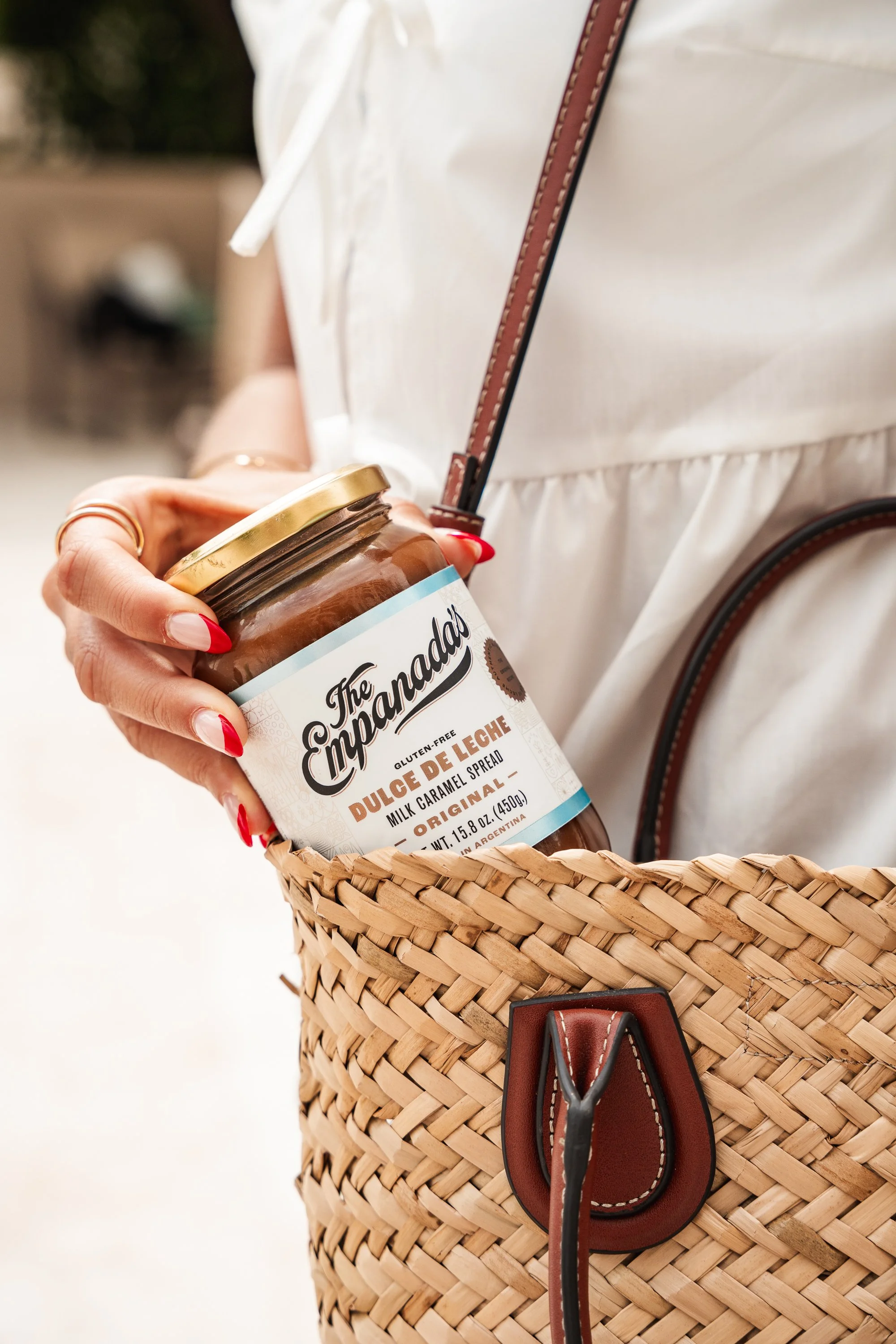A person holding a jar of dulce de leche in a woven basket, wearing a white blouse, with red painted nails, and a brown leather strap purse.