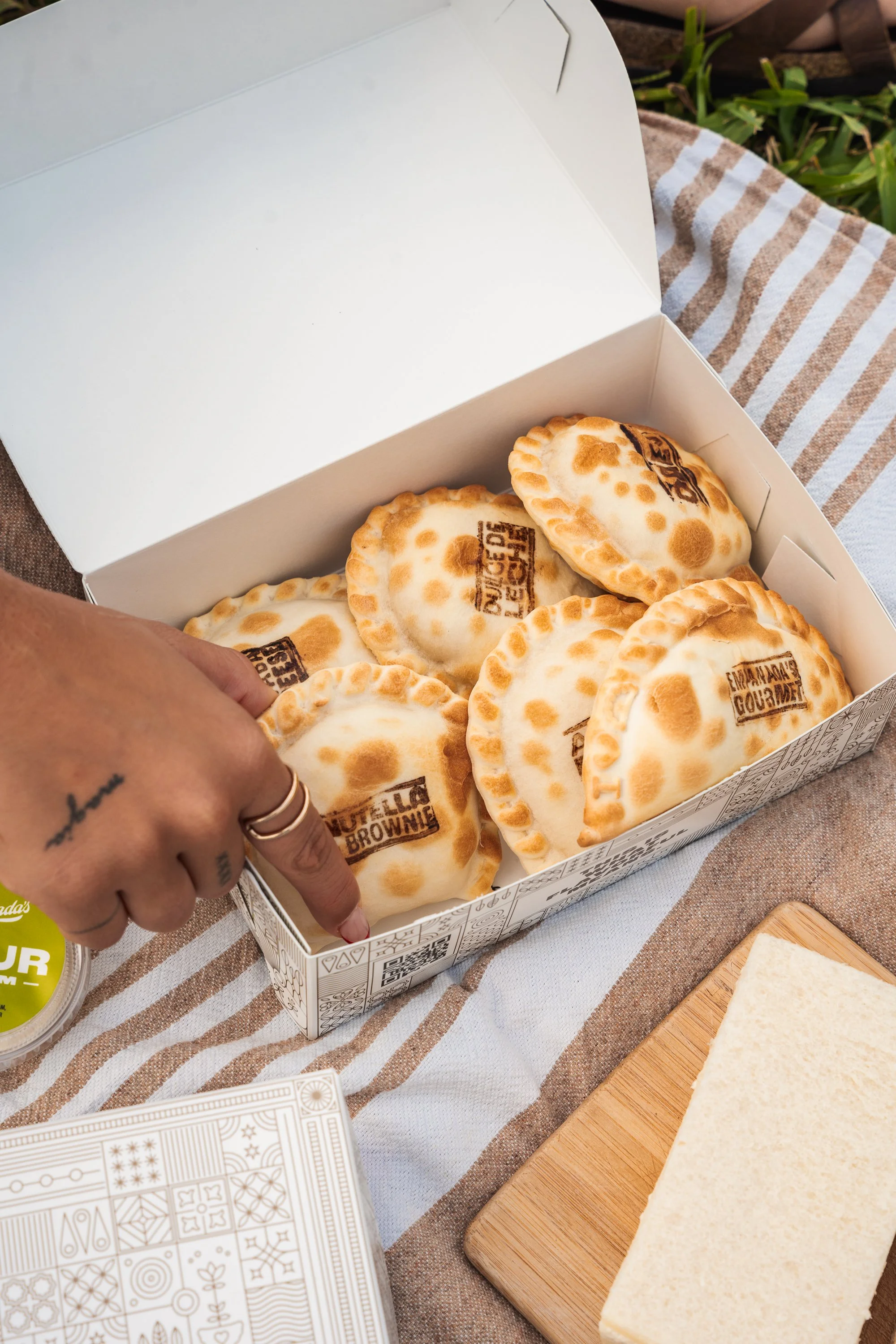 Open white box containing baked empanadas on a checkered fabric surface, with a person's hand pointing to one of the empanadas. There is a piece of sliced bread on a wooden cutting board nearby.