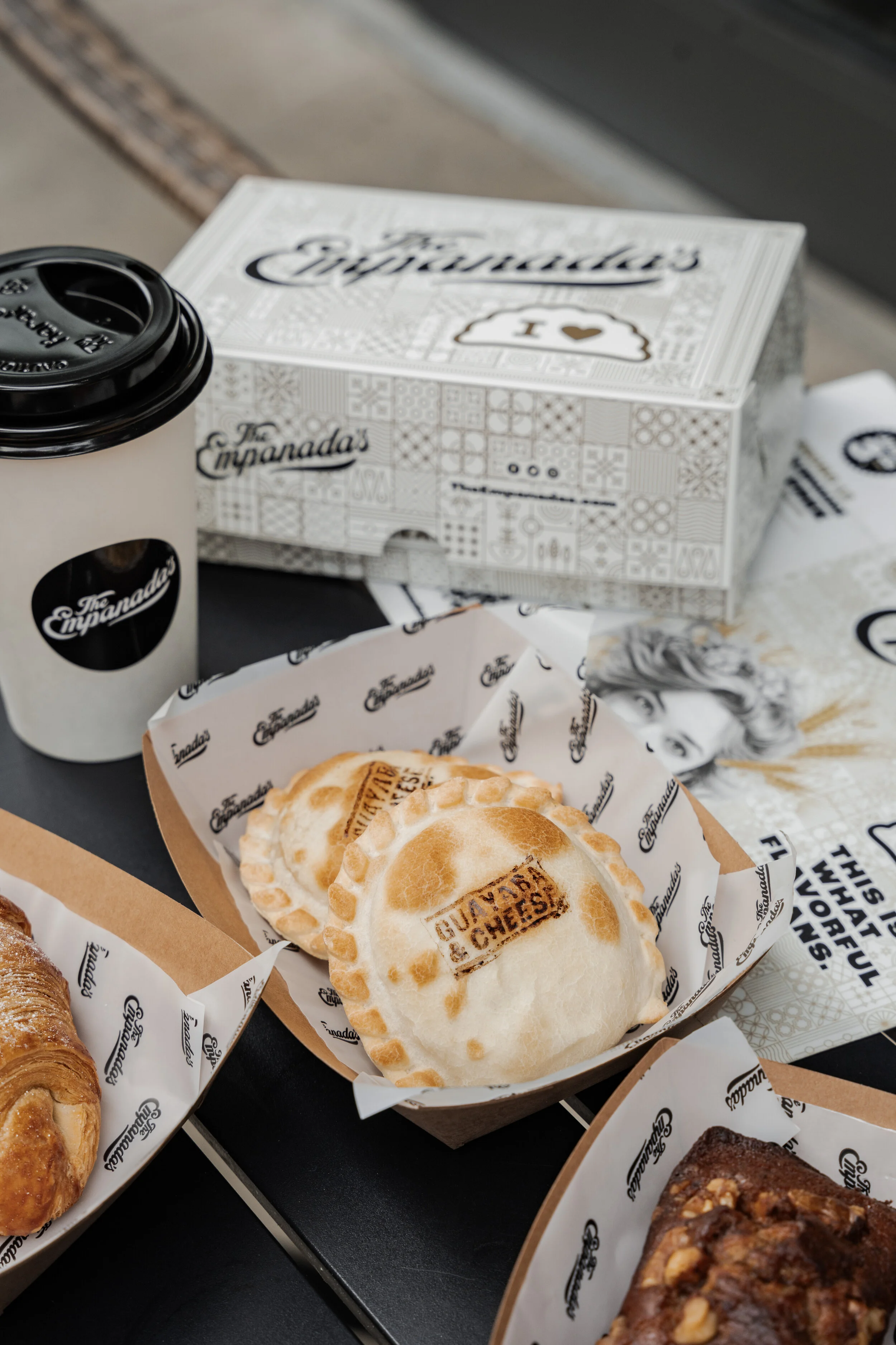 A tray with baked empanadas in branded paper liners and a cup of coffee on a black surface.