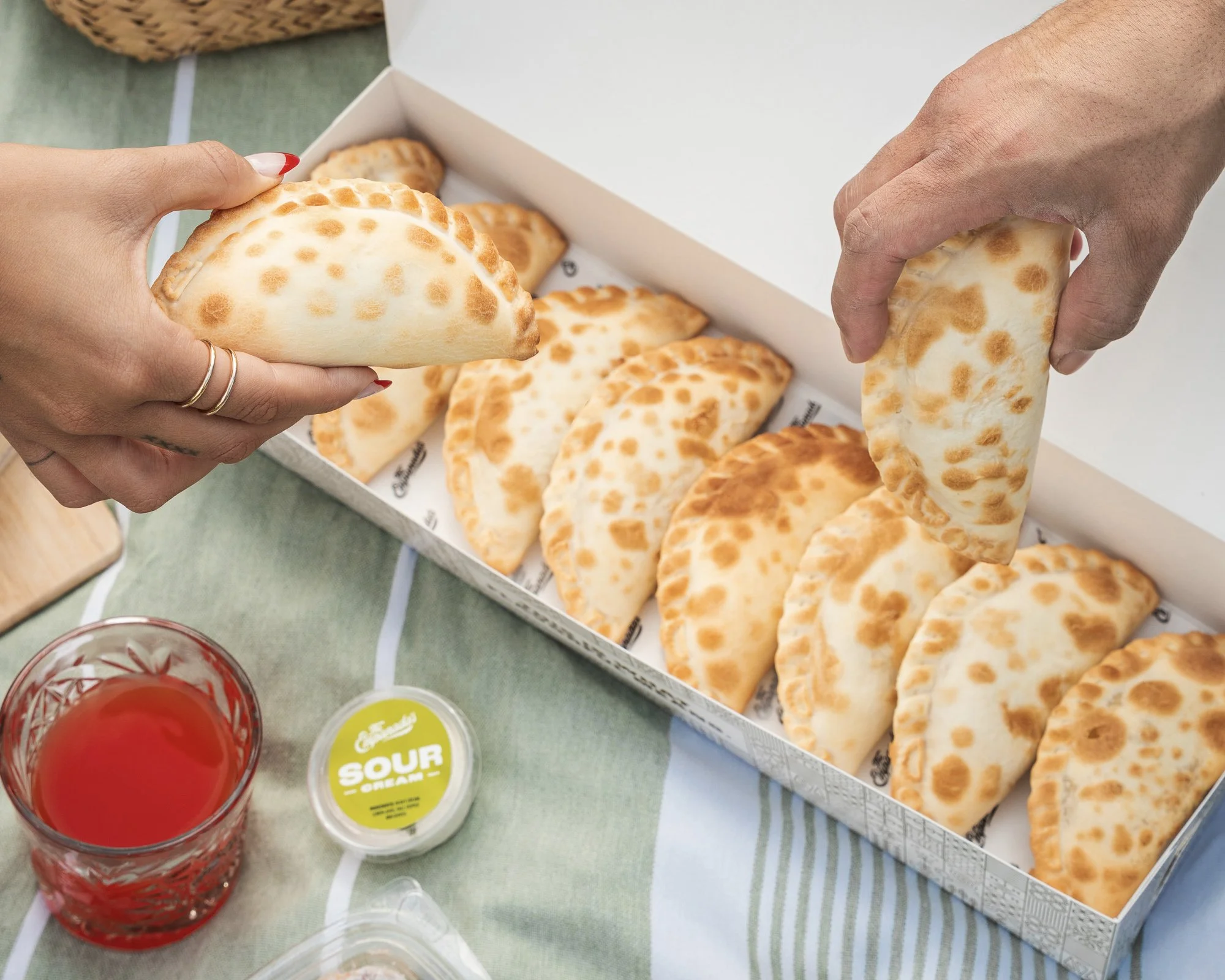 A person holding a baked empanada with a golden, slightly browned crust above a white box filled with more empanadas. There is a glass of red drink and a small container of sour cream nearby on a green tablecloth.
