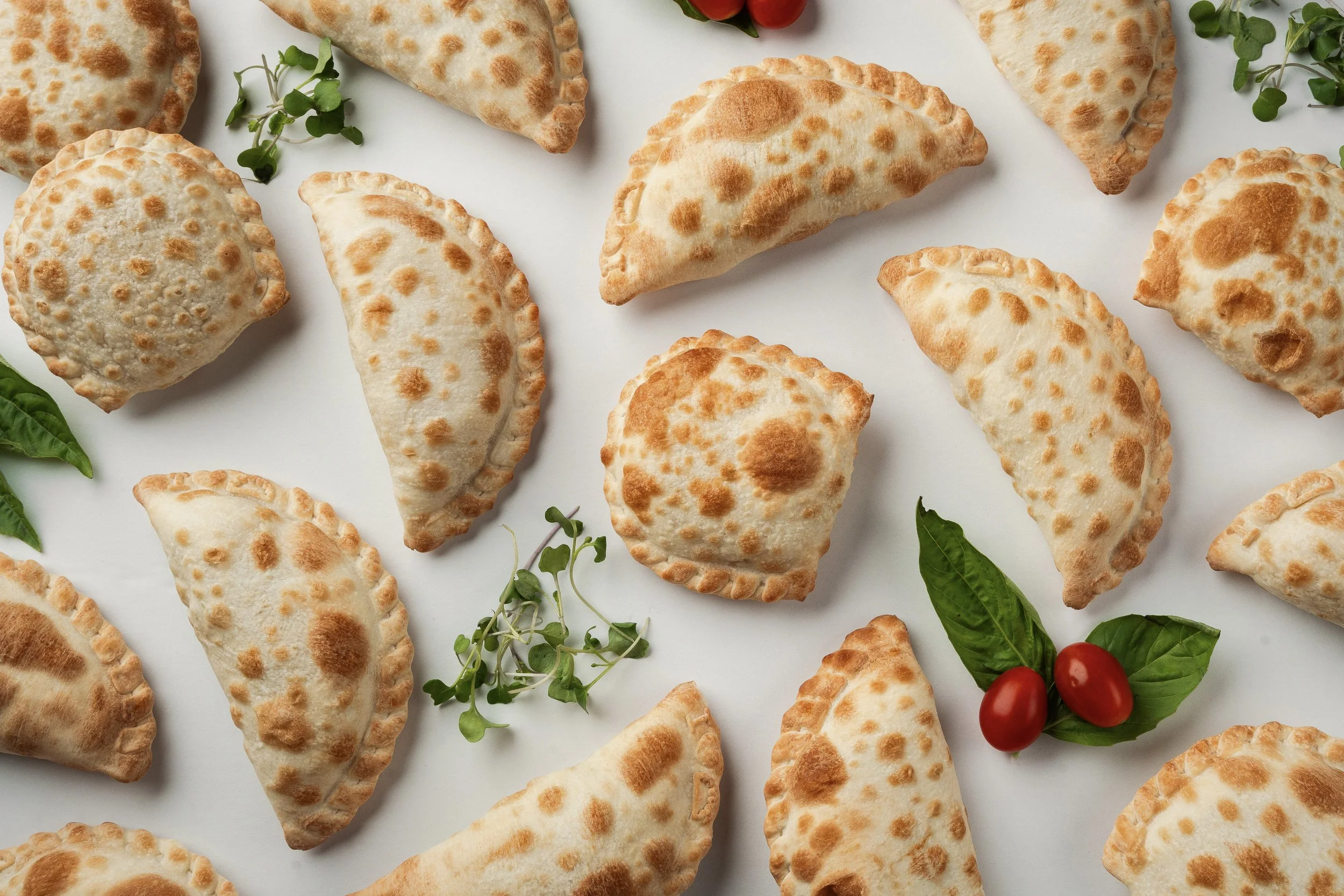 Assorted empanadas with a golden-brown crust, garnished with green herbs and small red tomatoes on a white surface.