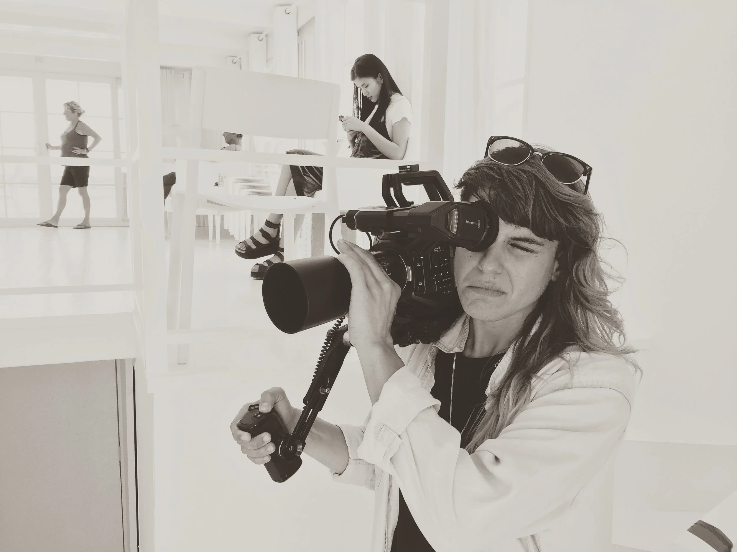 In the foreground, Zusel Escriba with long hair and sunglasses on their head is holding a video camera, filming the room.