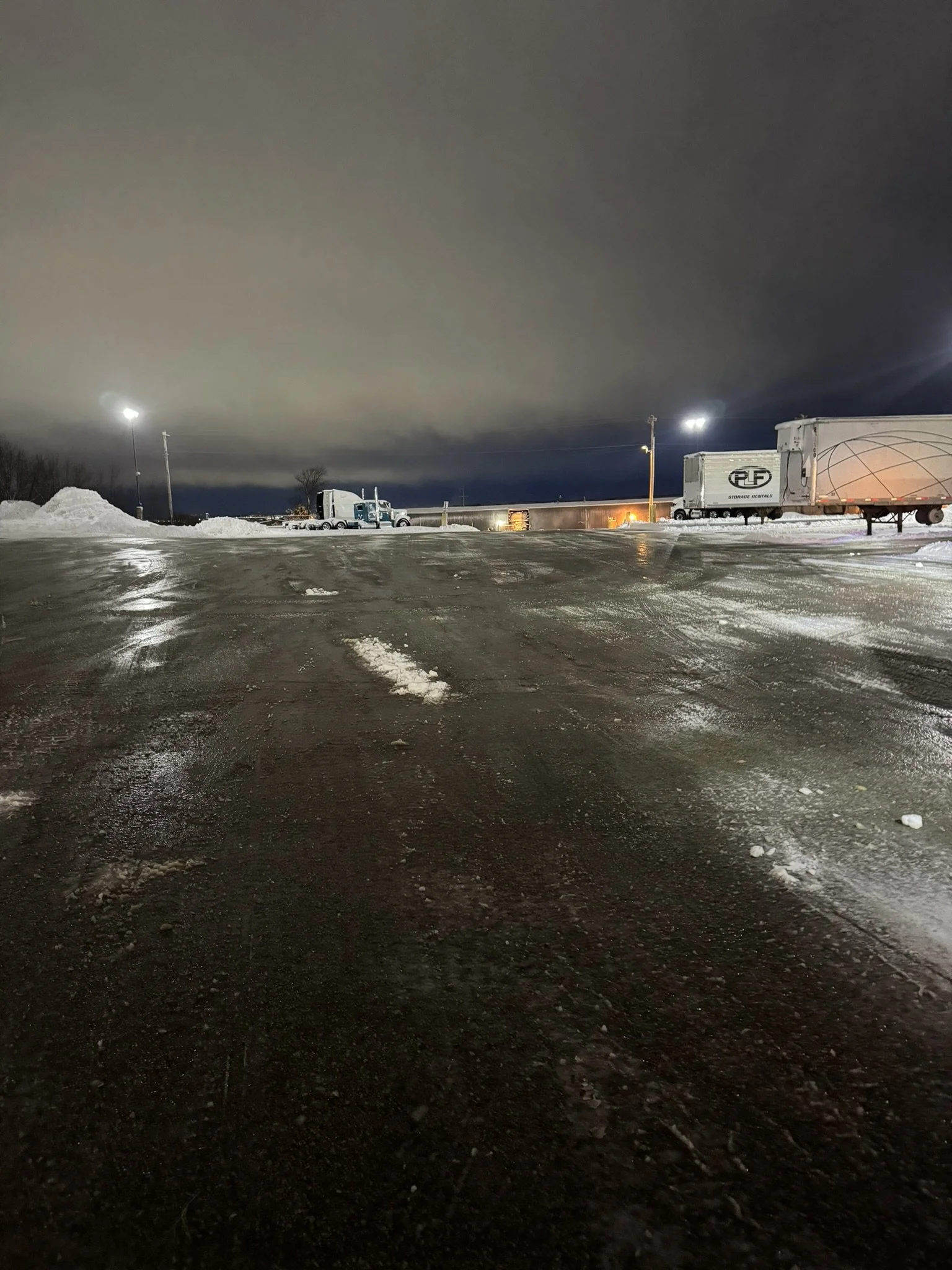 Nighttime scene of a parking lot with snow piles, several trucks, and trailer containers under illuminated tall lights, after using brine to melt the snow and ice.