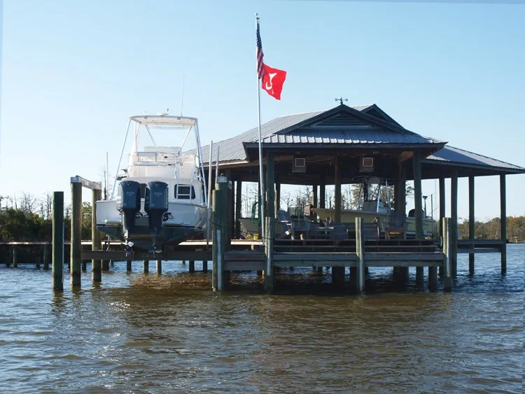 A boat docked at a covered pier on a body of water, with an American flag and a red flag flying, and trees in the background.
