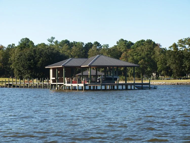 A wooden pier leading to a boat house with a metal roof on a calm lake, surrounded by trees under a clear sky.