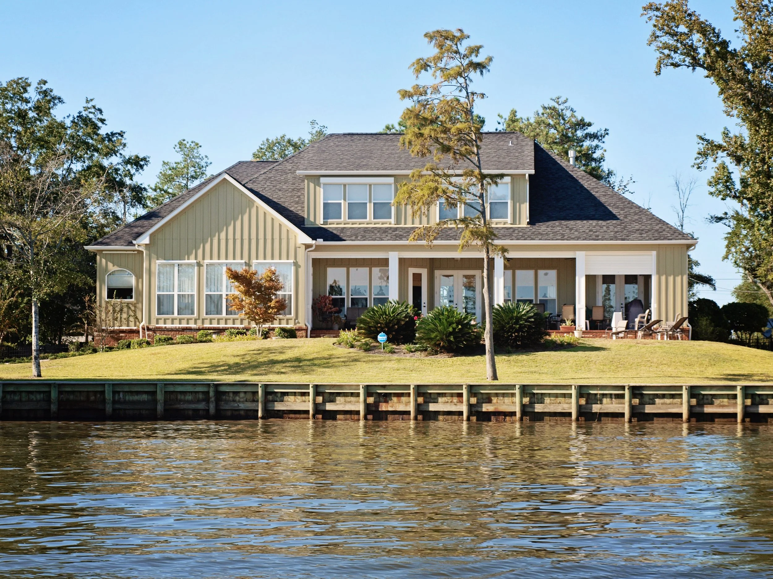 A house with a yellow exterior and a dark shingled roof, situated by a body of water with a wooden dock in the foreground and trees around it, under a clear blue sky.