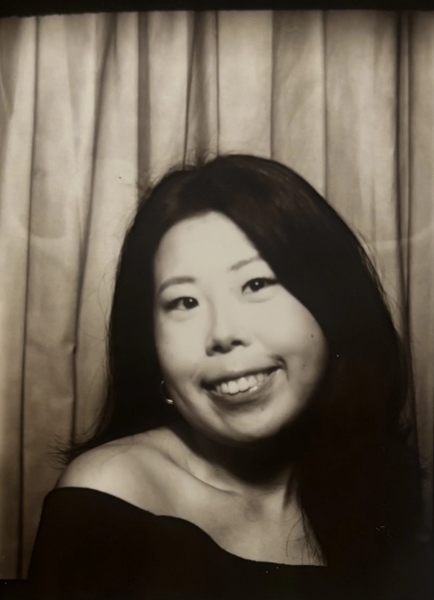 A black and white photo of a woman with long dark hair, smiling brightly, with a curtain in the background.
