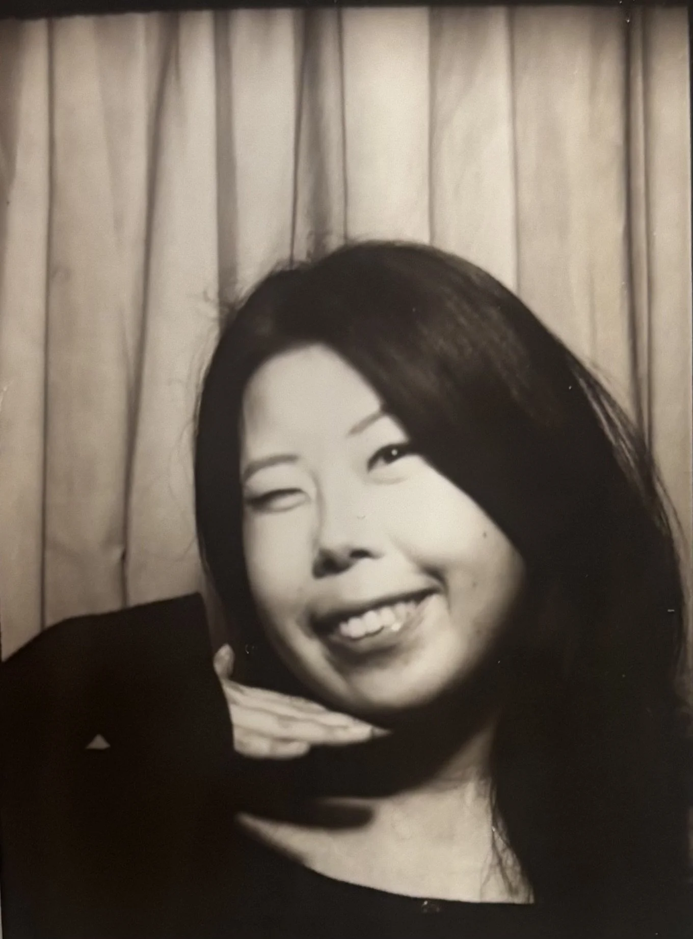 A black and white photo of a woman with long dark hair, smiling brightly, with a curtain in the background.
